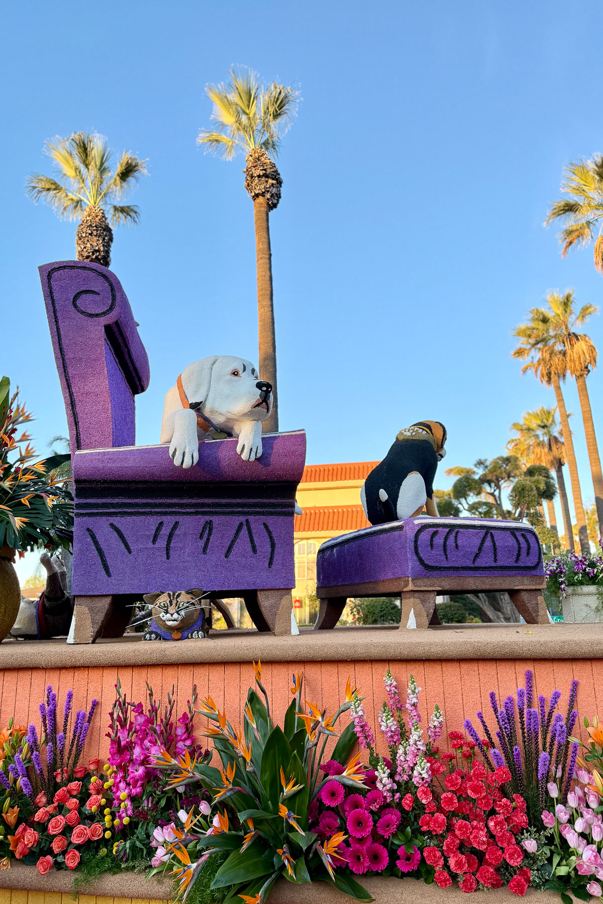 Colorful parade float with two beagle sculptures. One beagle faces forward on a purple chair; the other looks away on a matching ottoman. Bright flowers surround the float. Tall palm trees and a clear blue sky form the background.