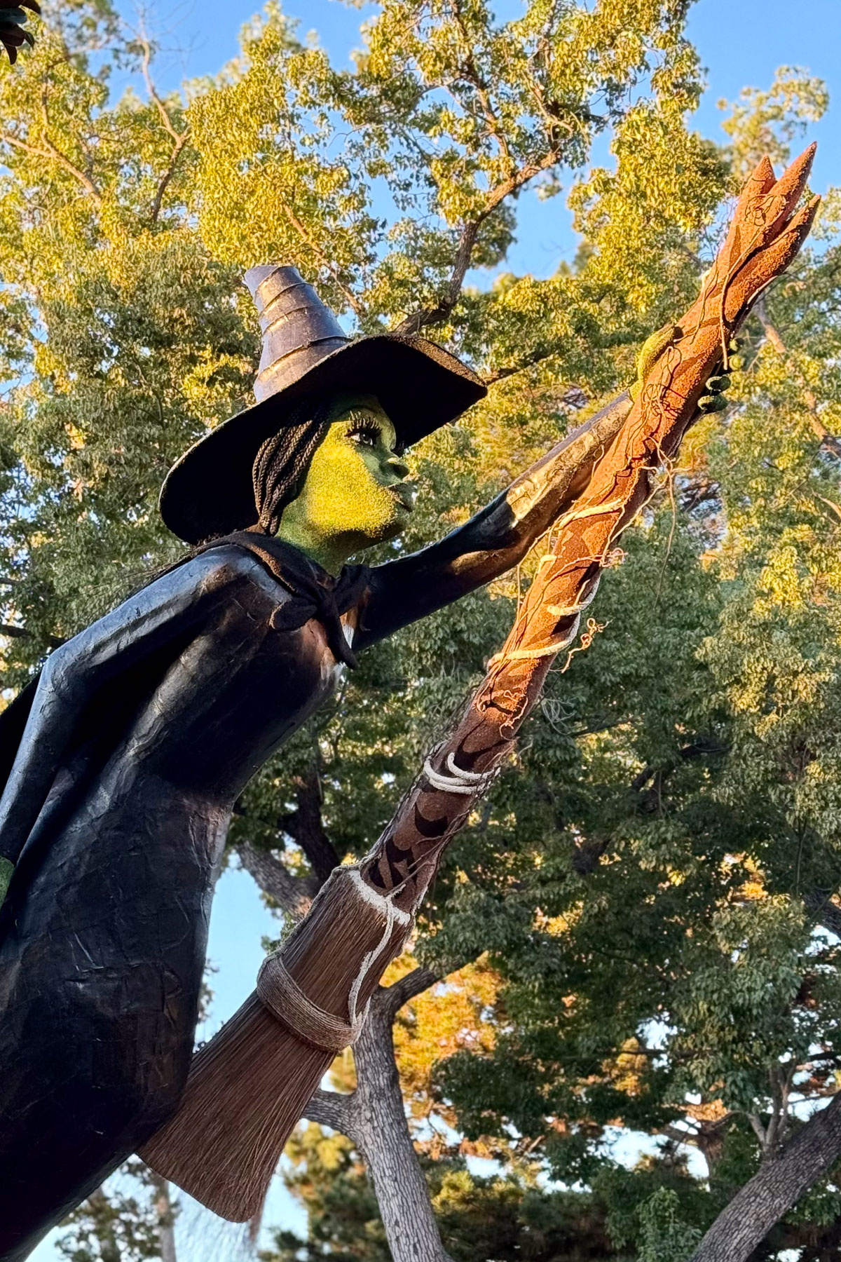 A sculpture of a green-faced witch wearing a black hat and cloak, holding a broomstick. The broomstick has decorative details and extends upwards. The background features trees with green foliage under a blue sky.