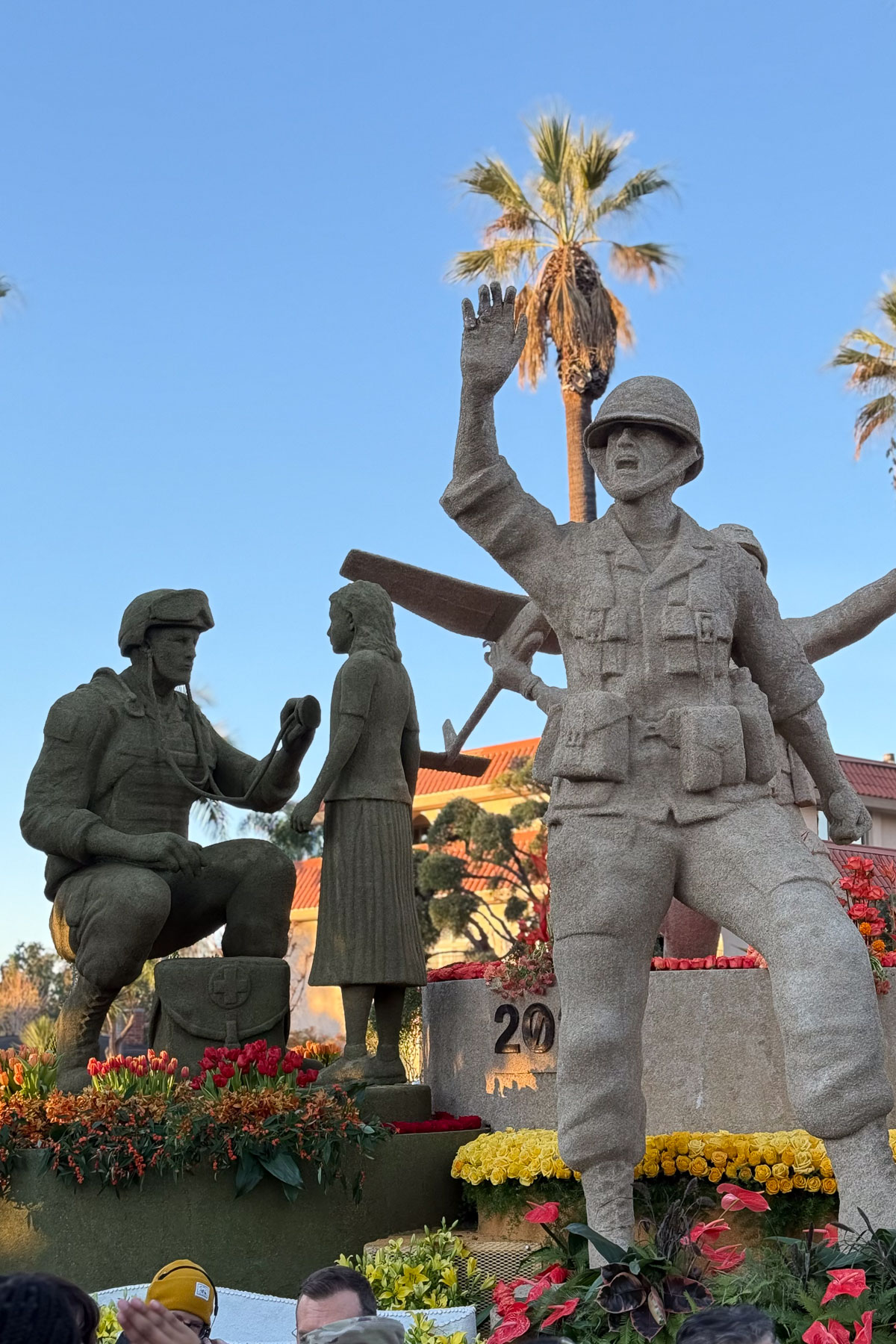 A sand sculpture depicting two soldiers in uniform, one kneeling and interacting with a civilian woman, while the other stands saluting. Surrounding them are vibrant flowers. Palm trees and a clear blue sky form the backdrop.