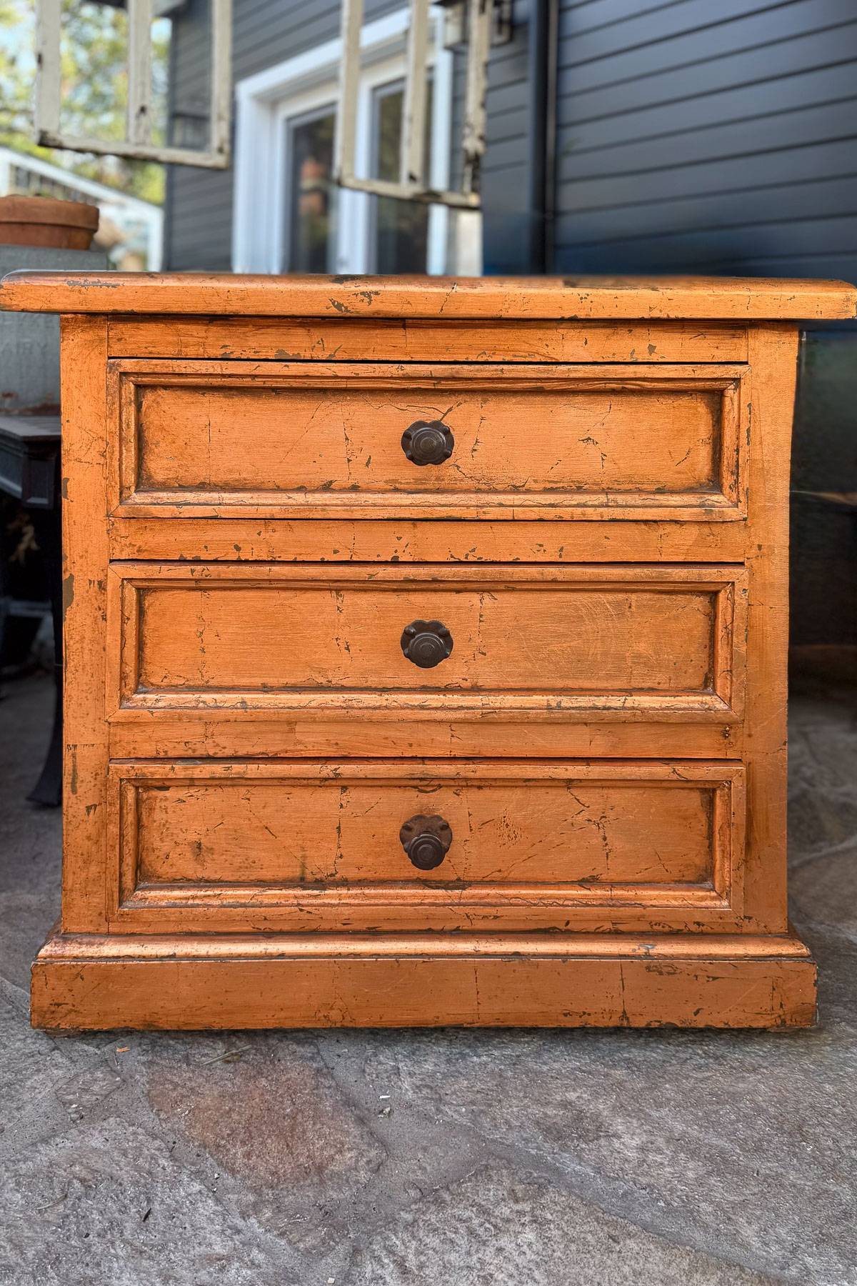 An orange distressed wooden chest with three closed drawers, each featuring a round, dark handle. The chest is set on a stone surface with a blurred background of a gray building and a window.