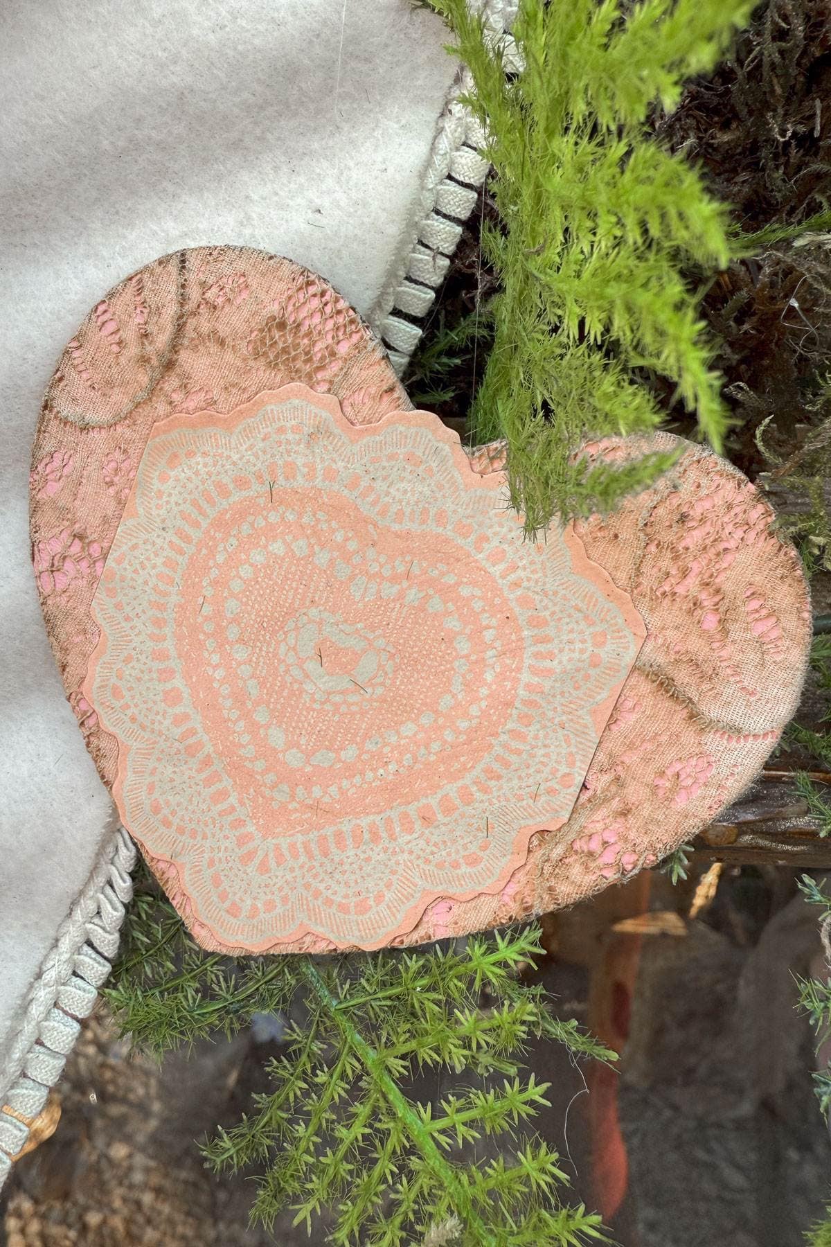 A heart-shaped decoration with intricate lace patterns in soft pink and beige hues is nestled among vibrant green foliage and a textured fabric in the background. The natural elements add a rustic and cozy feel to the scene.