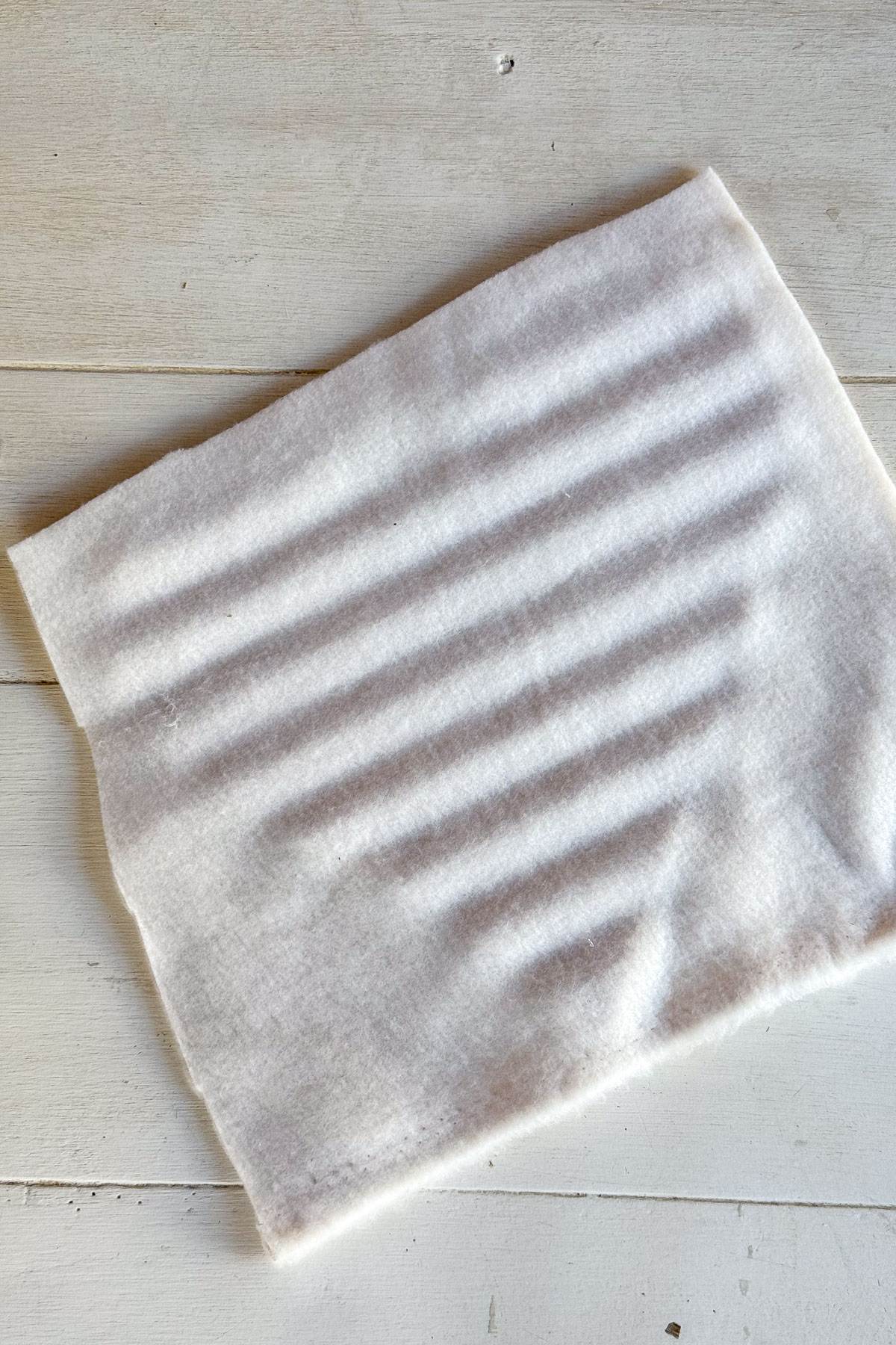 A piece of white fabric lies flat on a light wooden surface. The fabric has subtle horizontal ridges running across it, creating a softly textured appearance. The edges of the fabric are slightly uneven.