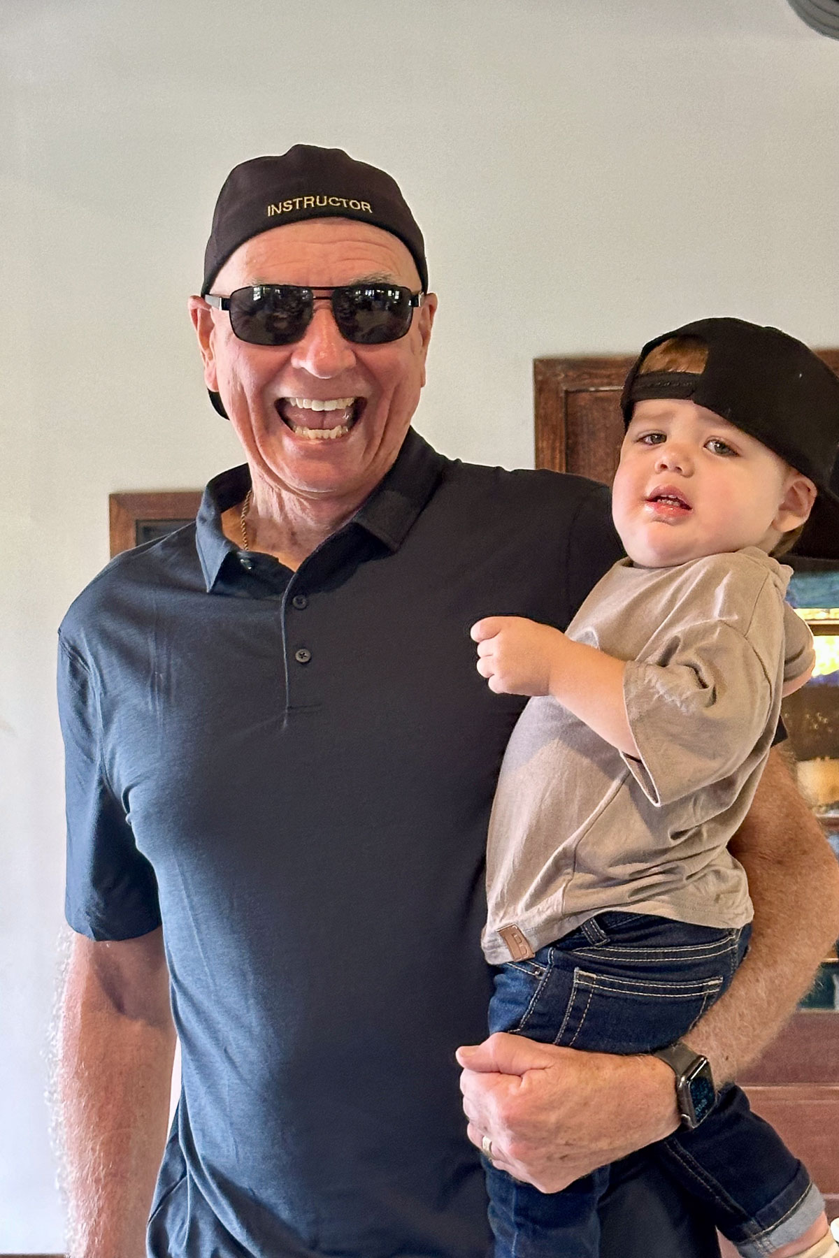 A smiling man wearing sunglasses and a cap labeled "INSTRUCTOR" holds a young child who is also wearing a backward cap. They are indoors and appear happy.