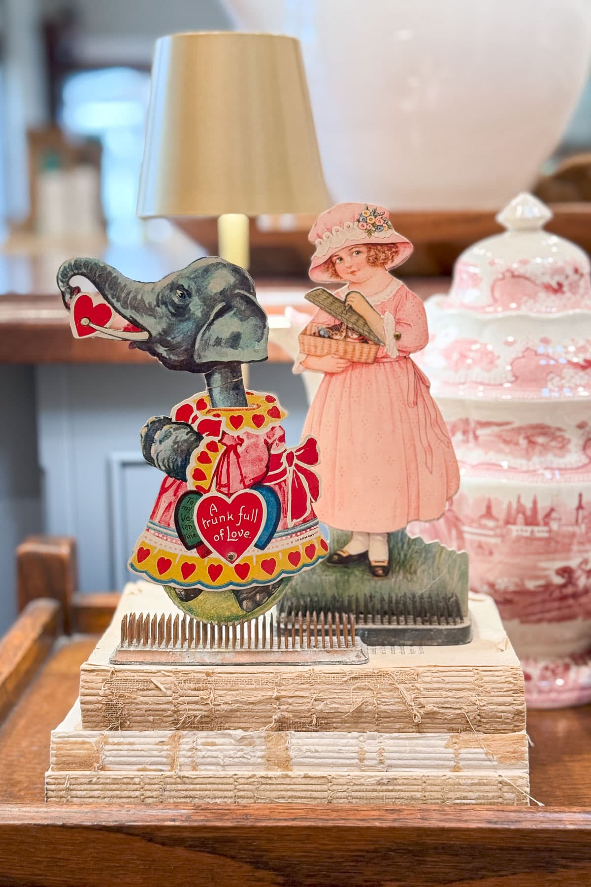 A vintage-style cardboard cutout of an elephant dressed in red and a girl in a pink dress holding a basket. They stand on a stack of books, with a red-patterned teapot in the background.