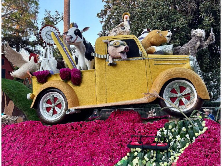 A colorful parade float features a yellow vintage truck adorned with vibrant flowers. Amongst the lush pink and white floral arrangements, several animated animal figures, including a cow and a pig, bring to mind a whimsical scene from someone’s "She Shed" on Sunday Sharing.