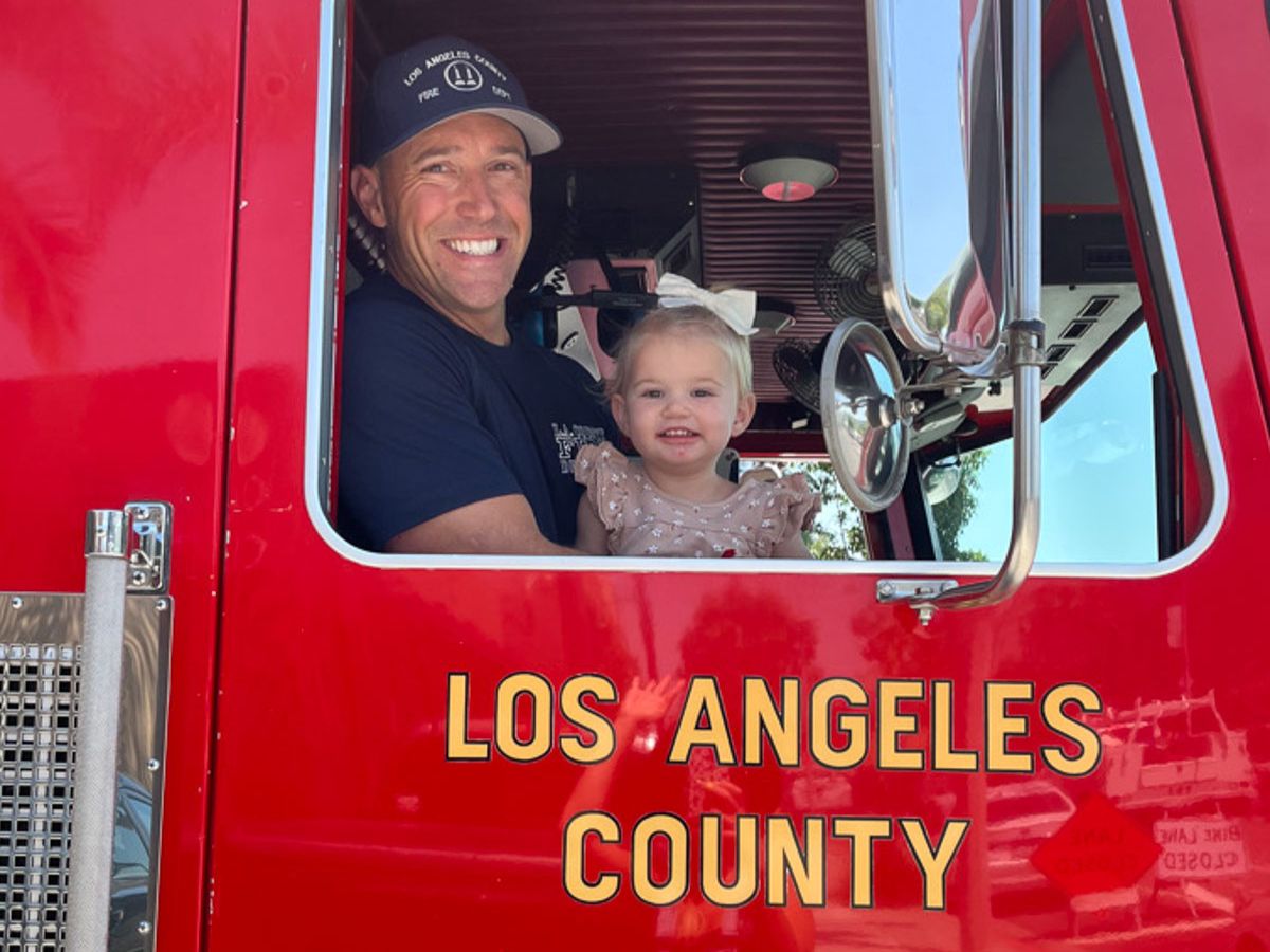 A smiling person in a cap and a young child, dressed in a light-colored dress with a bow in their hair, enjoy sitting together inside a bright red fire truck marked "Los Angeles County." It's the perfect moment for Sunday Sharing.