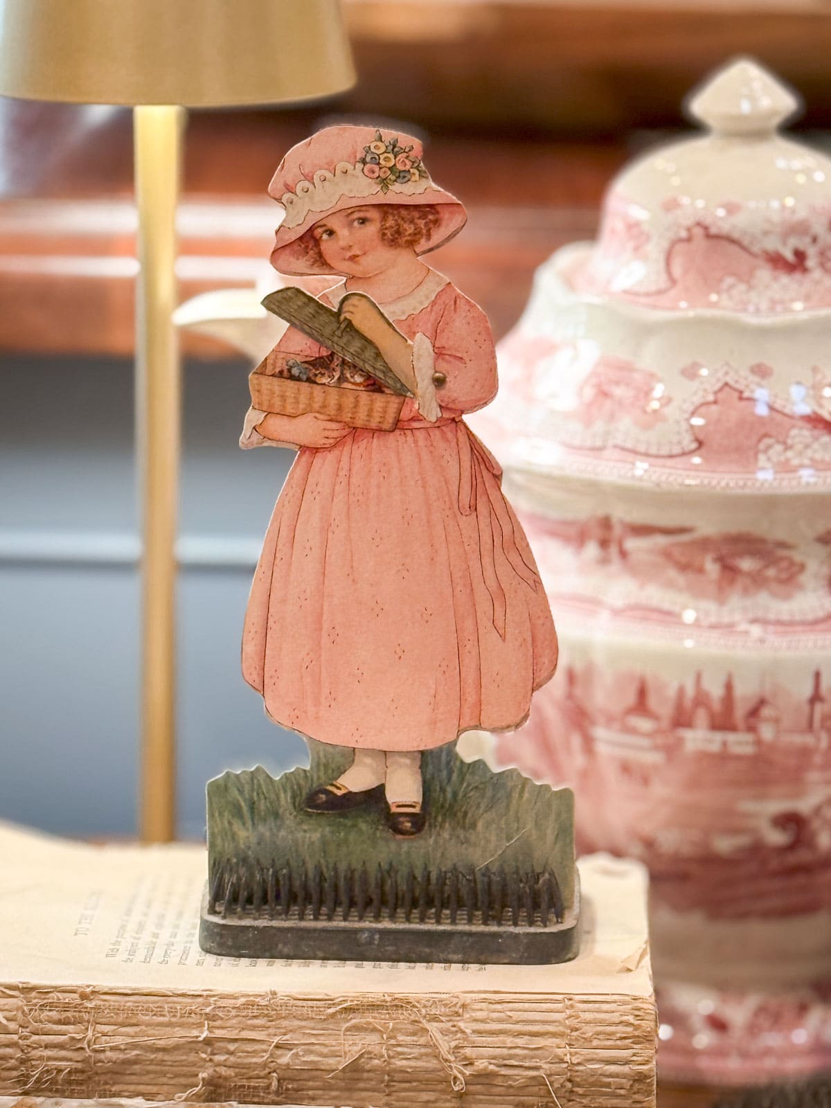 A vintage-style cardboard cutout of a young girl in a pink dress and bonnet holding a basket of bread stands on a stack of books. A decorative pink and white ceramic jar with a lid is in the background.