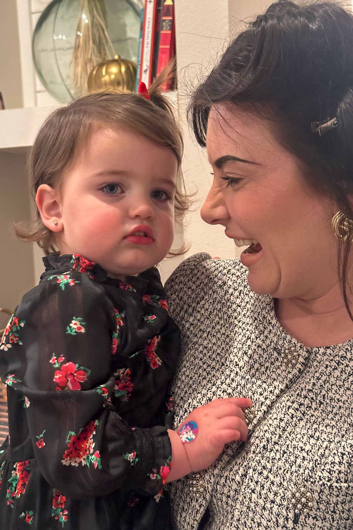 A woman smiling and holding a young child wearing a floral dress. The child has small earrings and a band-aid on their hand. They are indoors with a shelf and some decorations in the background.