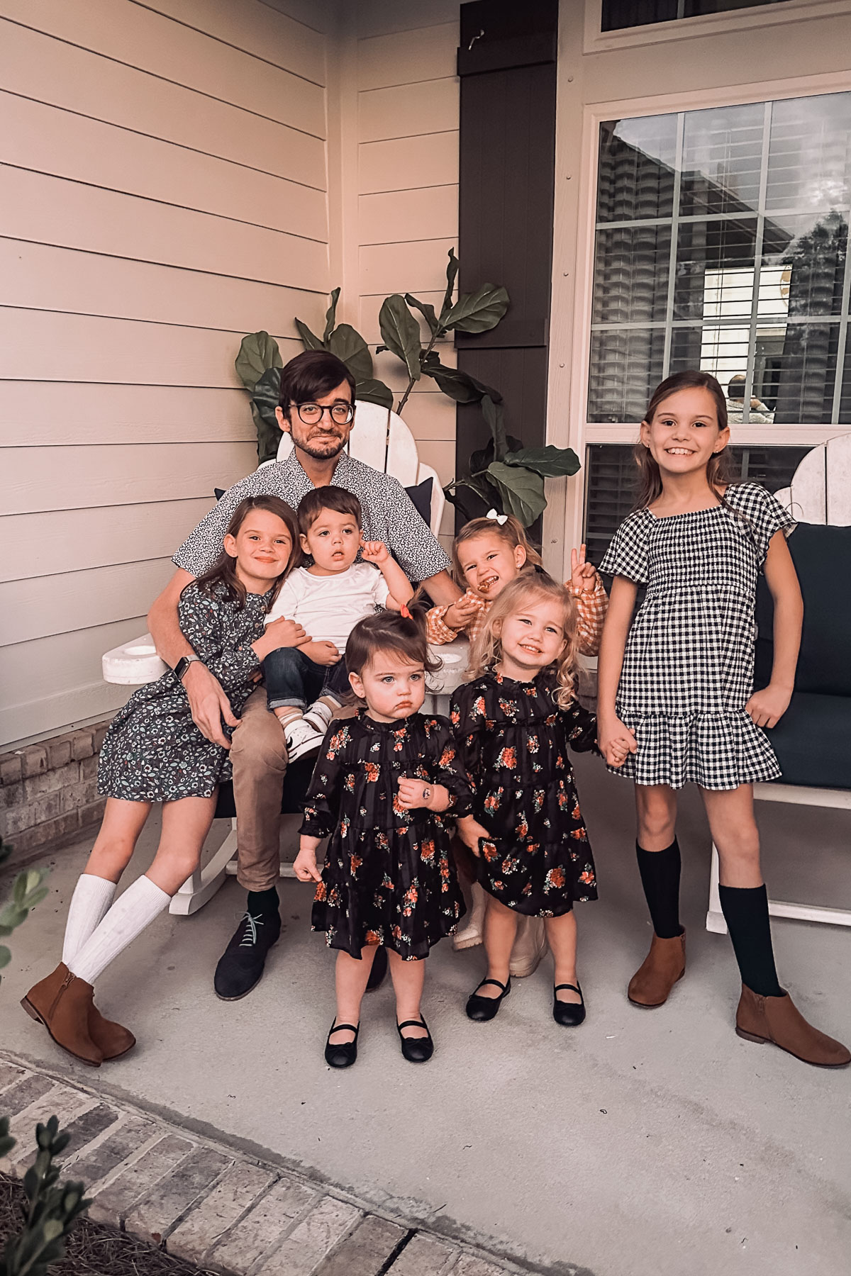 A man sits on a porch bench surrounded by six children. The children are dressed in various patterned outfits, with some standing and others sitting on his lap. A potted plant and a window are in the background.