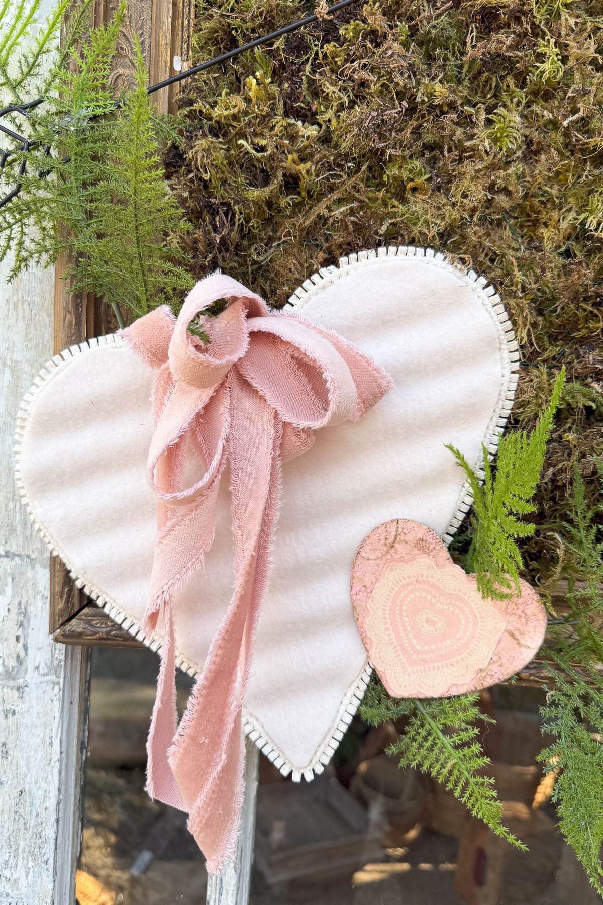 A decorative display featuring a large white heart with a pink ribbon bow attached, accompanied by a smaller pink heart. They are set against a backdrop of green ferns and moss on a rustic surface.