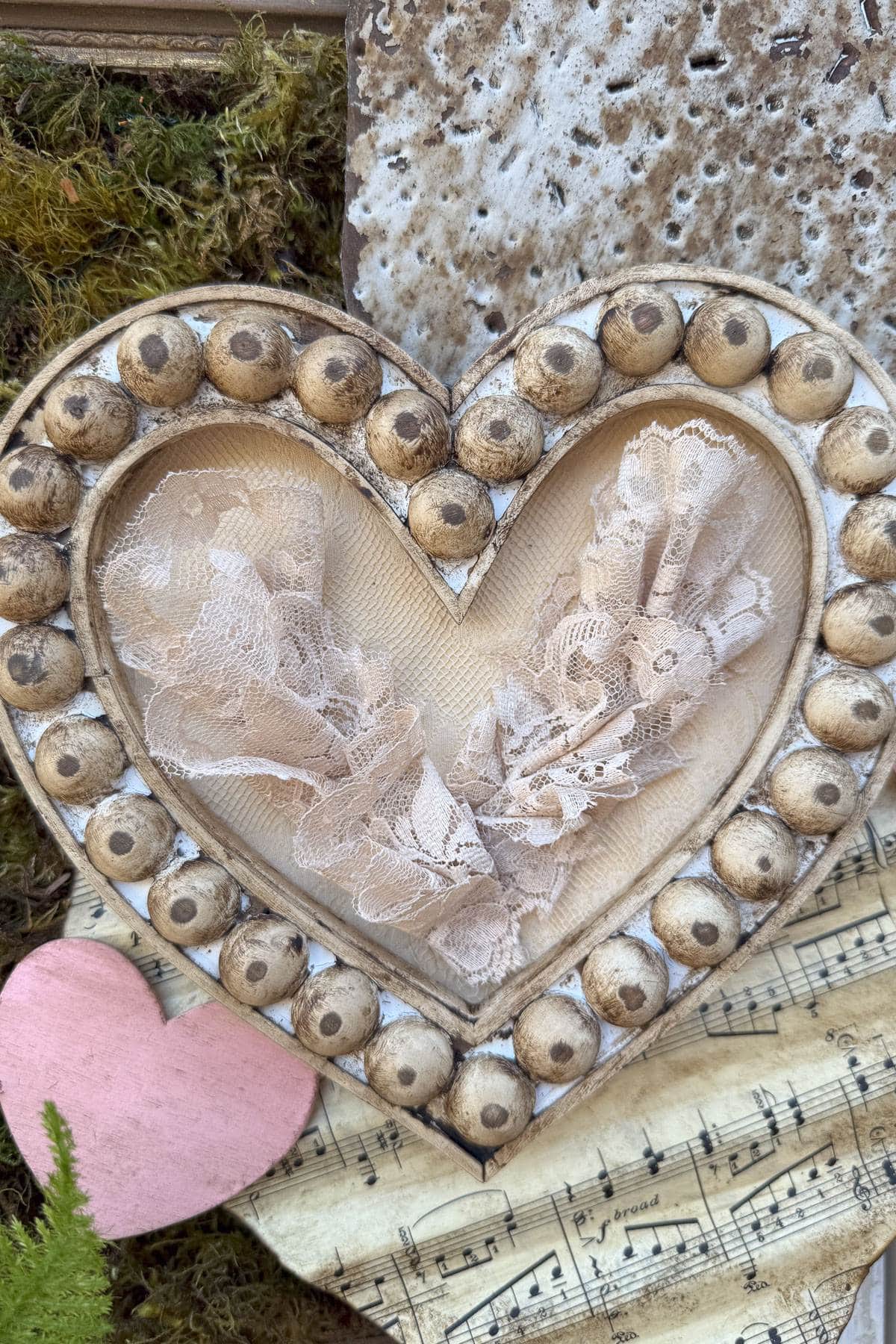 A wooden heart frame decorated with round beads, showcasing delicate lace inside. It's placed on sheet music with a small pink paper heart and surrounded by moss, evoking a rustic and romantic atmosphere.