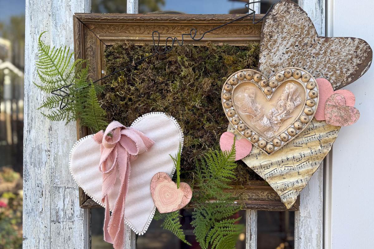 A rustic wall decoration features three hearts: a pink velvet one with a bow, a metallic one with an embossed design, and one made from sheet music. Moss, ferns, and a subtle "love" wire complete the arrangement, framed in old wood.