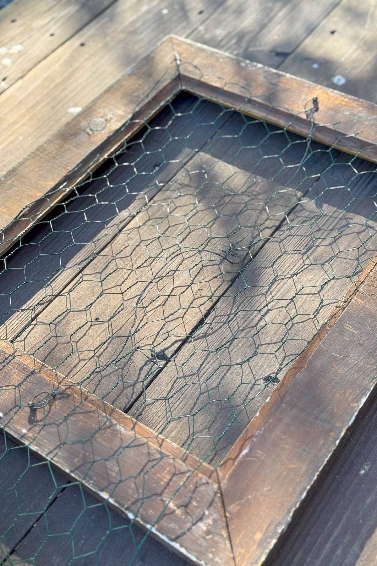 A weathered, rectangular wooden frame with rusted chicken wire laid over it, resting on a wooden deck. Sunlight casts soft shadows across the surface, highlighting the texture of the wood and wire.