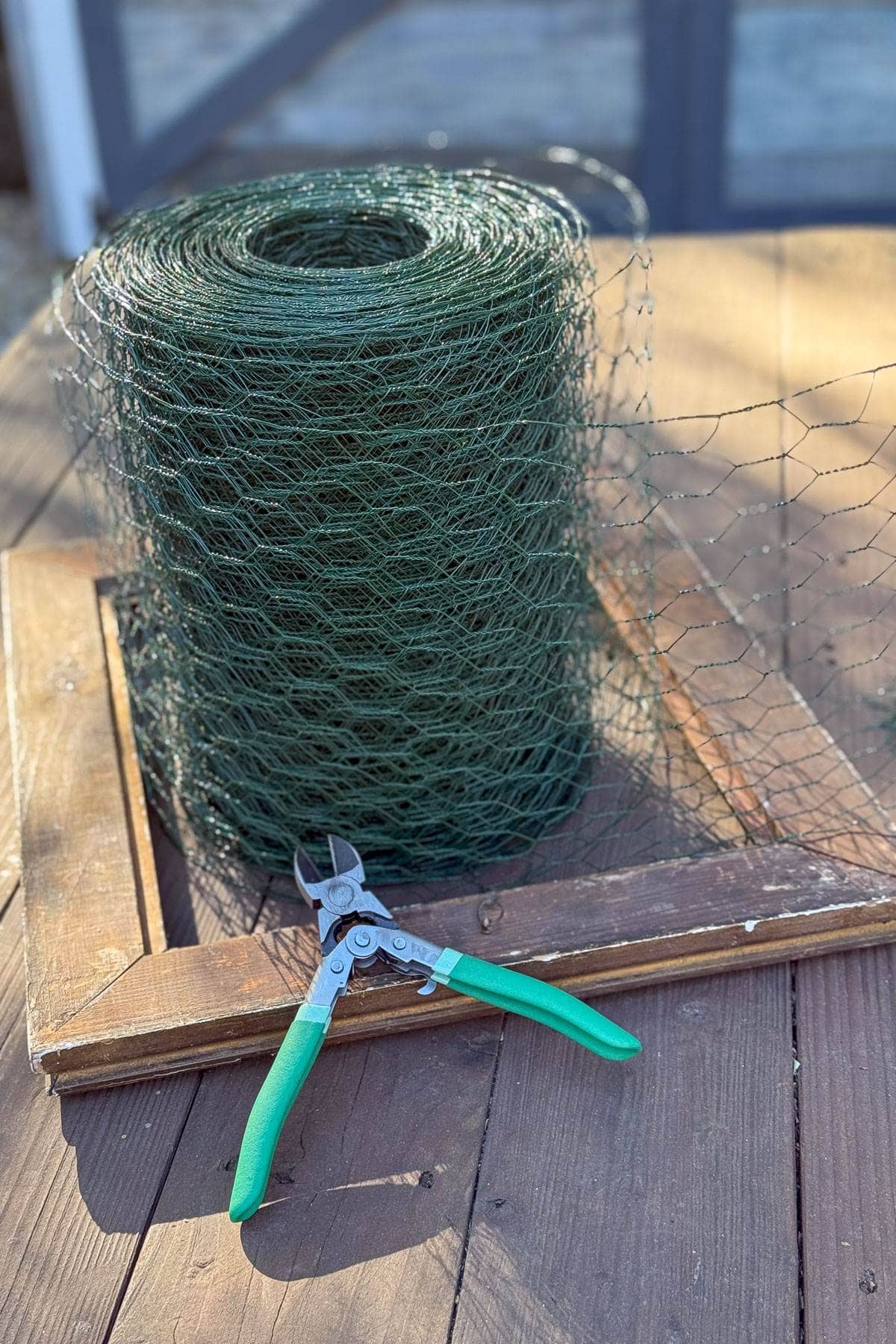 A roll of green wire mesh sits on a wooden surface, partially unrolled. A pair of wire cutters with green handles rests in front of it.