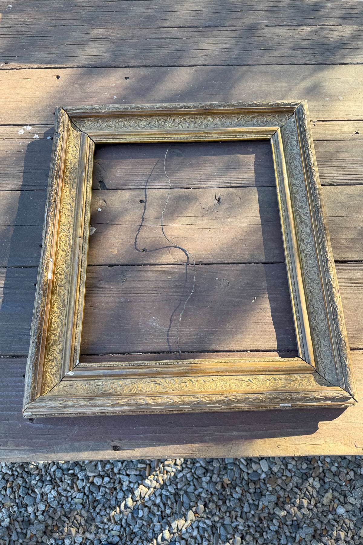 An empty, ornate, gold-colored picture frame lies on a wooden deck. The frame has intricate floral patterns. A faint line shadow resembling a face appears inside the frame. Pebbles can be seen on the ground beside the deck.