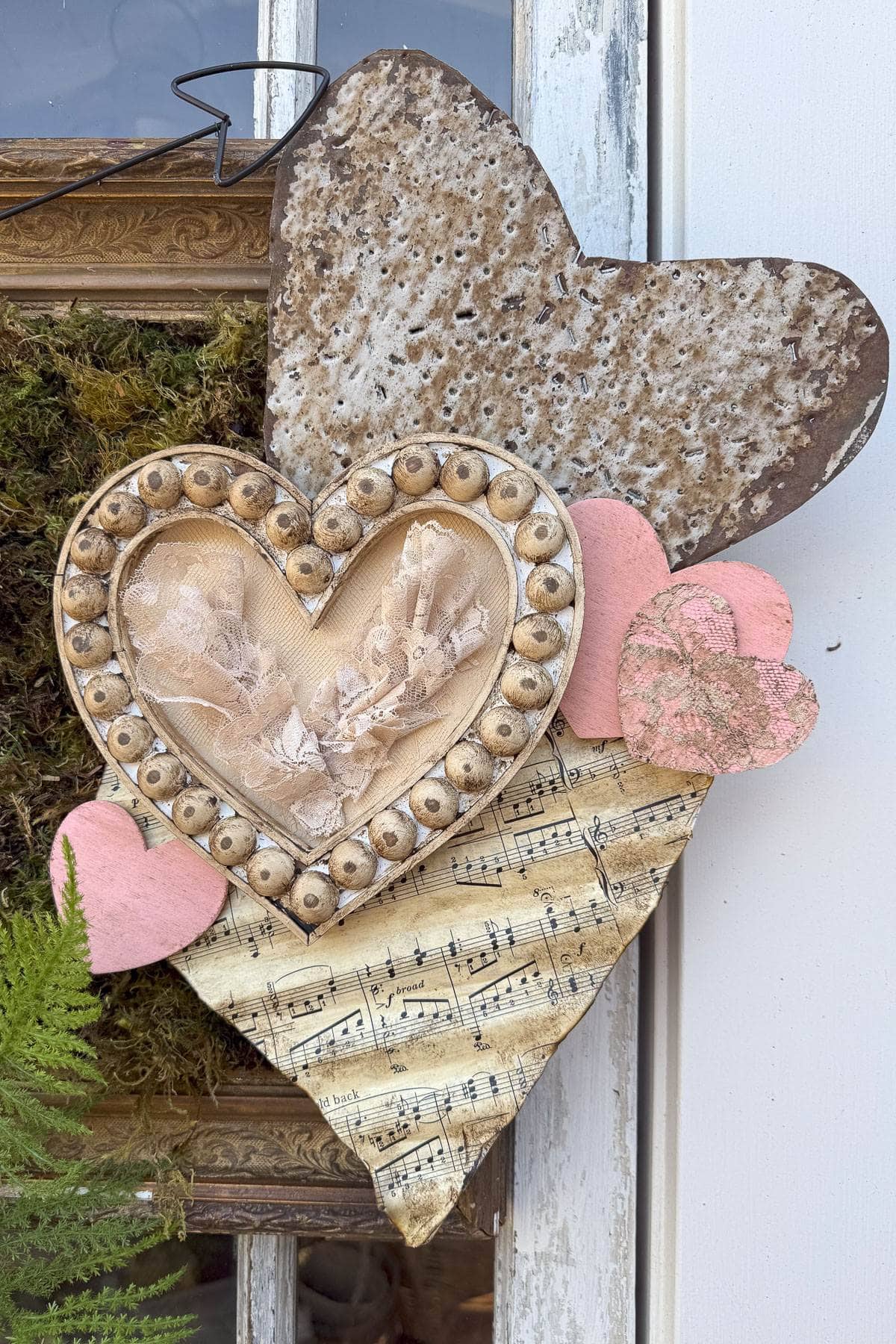 A decorative arrangement featuring layered hearts: a weathered metal heart, a lace-trimmed heart framed with beads, a heart covered in sheet music, and small pink hearts. They are creatively displayed against a rustic background with fern plants.