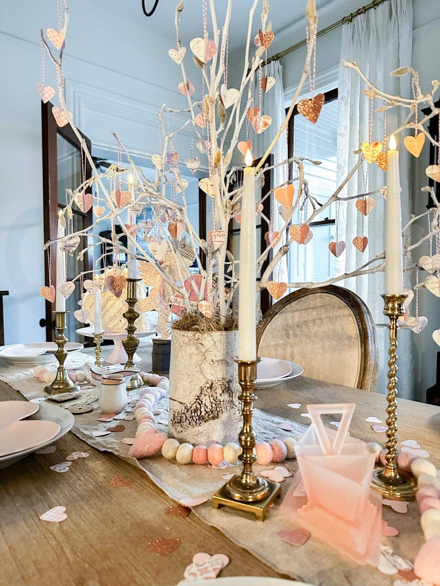 A wooden dining table decorated with heart garlands, pink and white ornaments, brass candlesticks, and a vase with white branches makes for charming Valentine's Day dinner table decoration ideas in a cozy, elegant room filled with soft natural light.