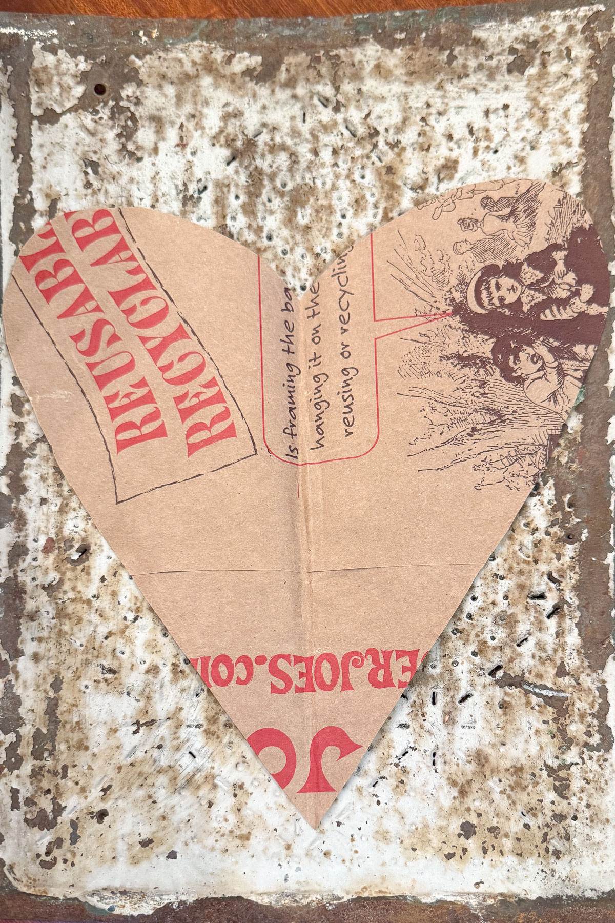 A heart-shaped piece of cardboard with red text saying "REUSABLE" and "RECYCLABLE" on a weathered, white-tiled surface. The cardboard features a sketch of people in rustic attire, surrounded by plants.