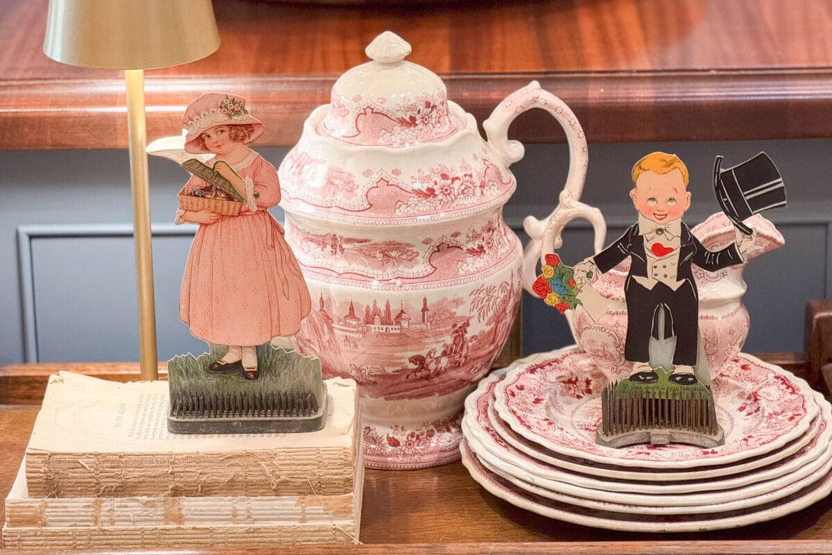 A decorative scene with a ceramic pitcher and plates featuring a pink pastoral design. In front, a vintage-style standing cutout of a girl in pink and a boy in a suit holding a bouquet and hat. A stack of books sits beneath a golden lamp.