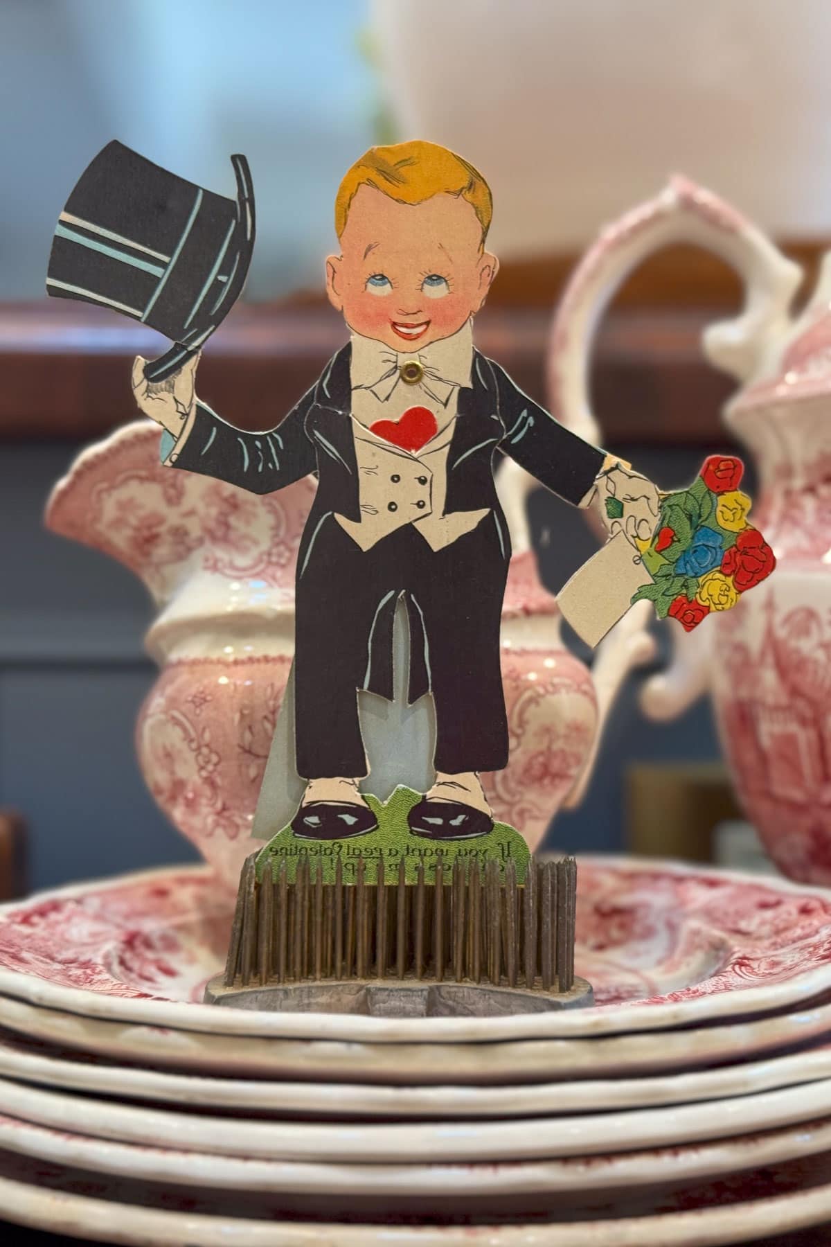 A vintage paper cutout of a smiling boy in a tuxedo holding a top hat and a bouquet of colorful flowers. He stands on a small platform. Behind him are plates and teapots with intricate red patterns.