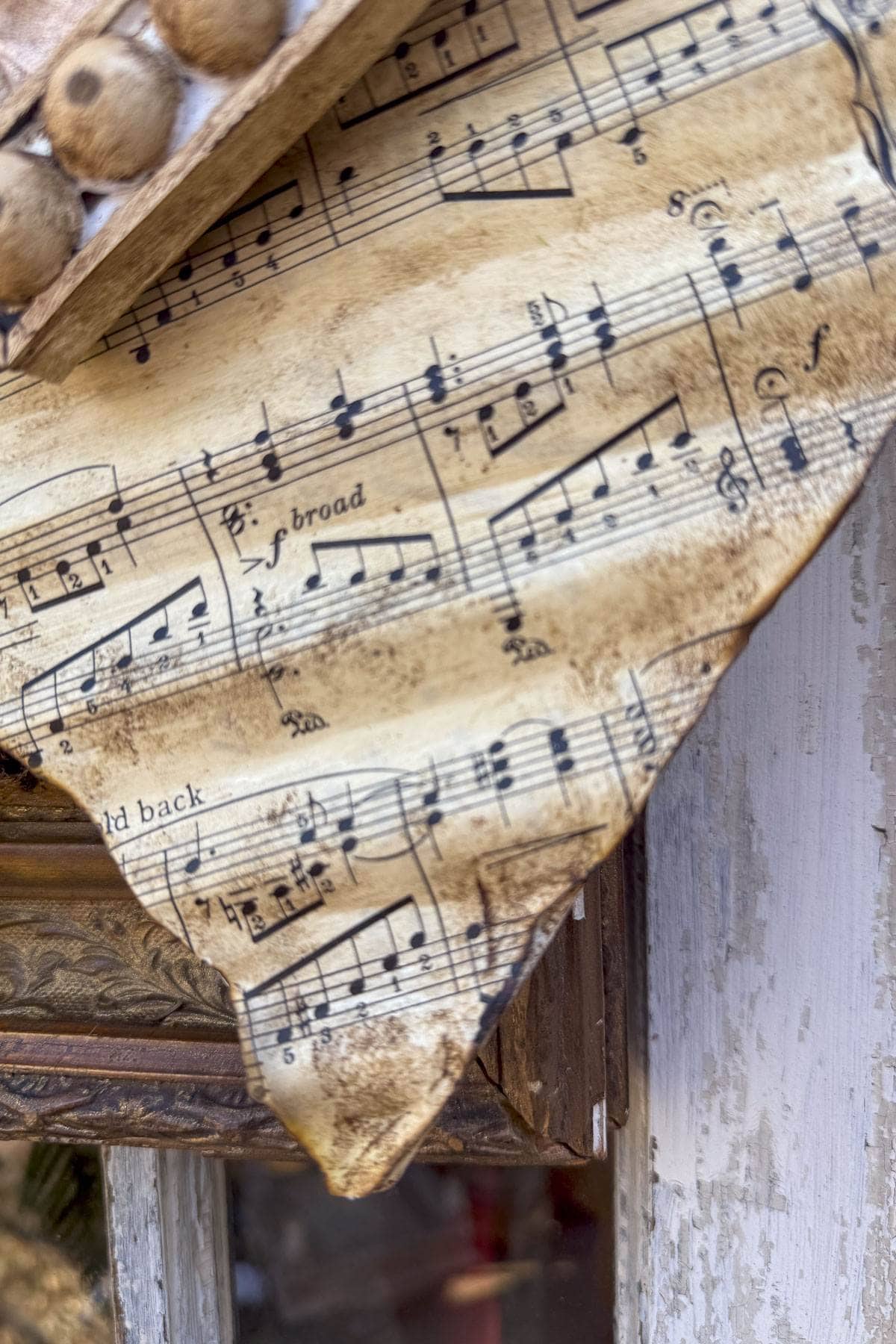 A close-up of a worn sheet of music with frayed edges. The paper shows musical notes and annotations, with a vintage, aged appearance. The background includes a blurred, rustic wooden surface.