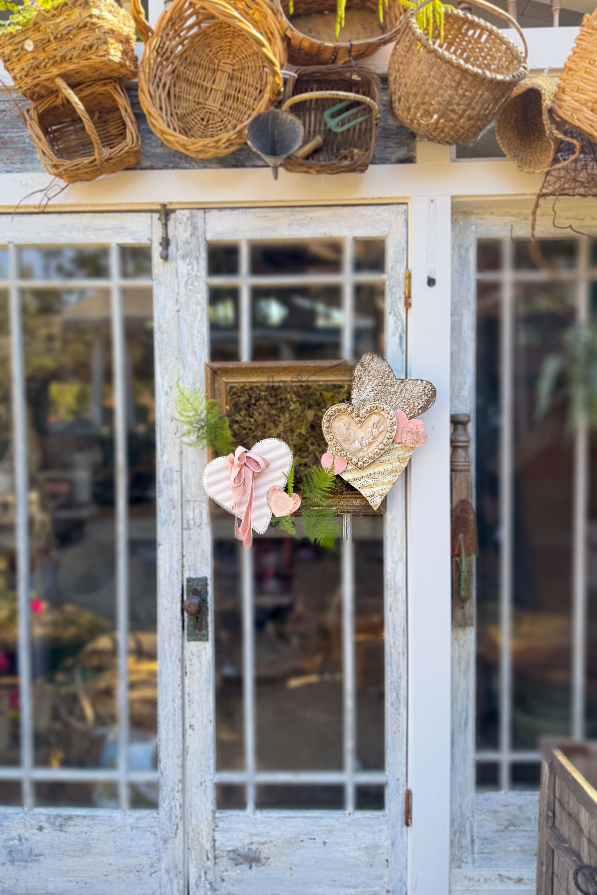 A rustic door with glass panes features a heart-shaped wreath, embodying the essence of Valentine decor. The wreath showcases fabric hearts, pink bows, and greenery. Baskets hang above the door, adding cozy DIY decorating vibes and unique vintage charm to the scene.