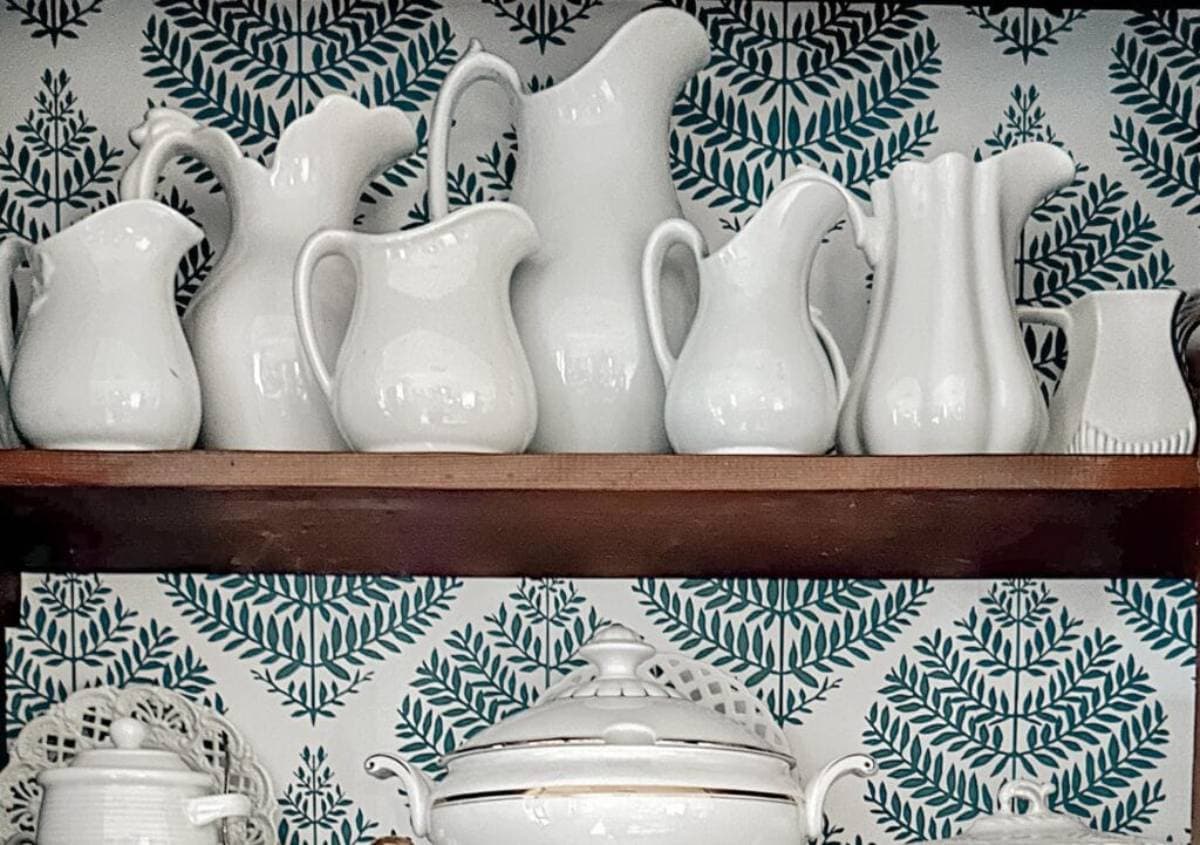 A collection of white ceramic pitchers is displayed on a wooden shelf against a blue fern-patterned wallpaper. Below the shelf, a white ceramic soup tureen with a lid adds to the elegant arrangement.