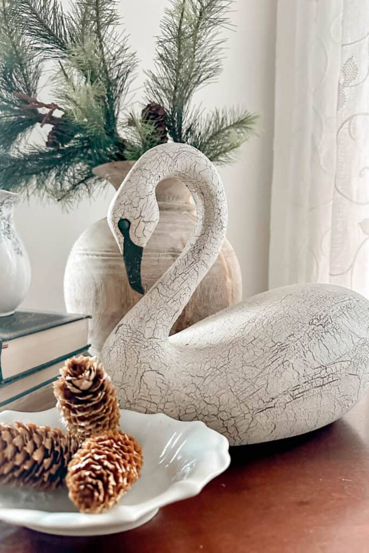 A decorative setup with a white crackle-textured swan sculpture on a table. Nearby, there are pinecones in a dish, stacked books, and a vase with green pine branches. The background features a soft, patterned curtain.