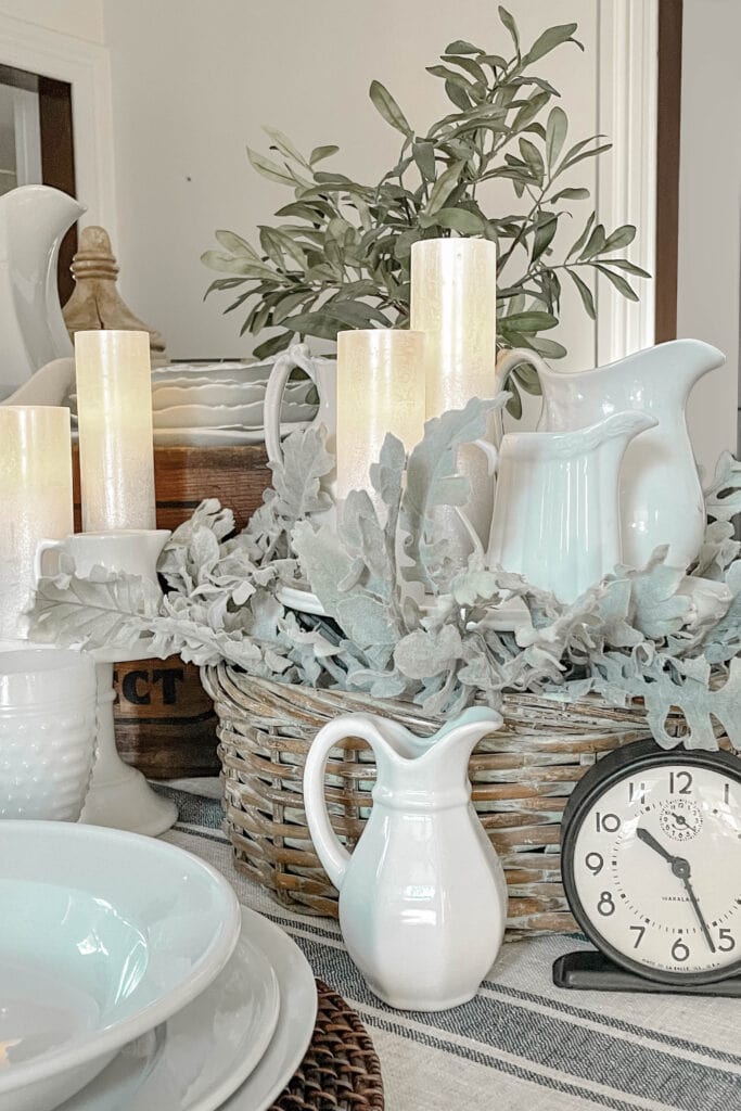 A dining table decorated with white ceramic dishes, pitchers, and glowing candles sets a cozy scene. Eucalyptus leaves in a wicker basket add freshness. A small clock displays the time, while the subtly striped tablecloth enhances the rustic charm of winter decorations not Christmas-focused.