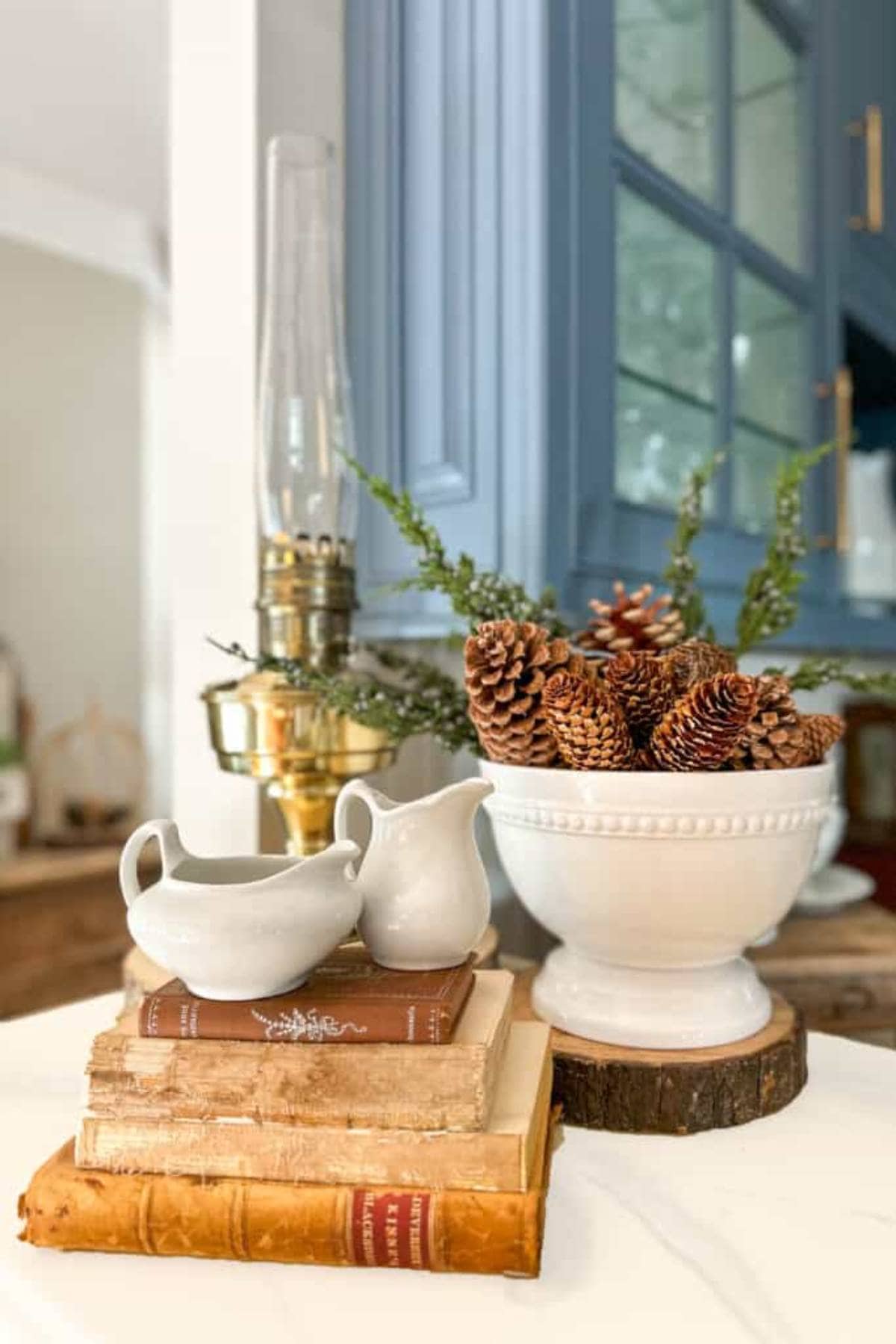 A cozy kitchen scene with a stack of vintage books topped by two white ceramic pitchers. A white bowl filled with pinecones and greenery sits nearby. An antique brass oil lamp adds a rustic touch, with blue cabinetry in the background.