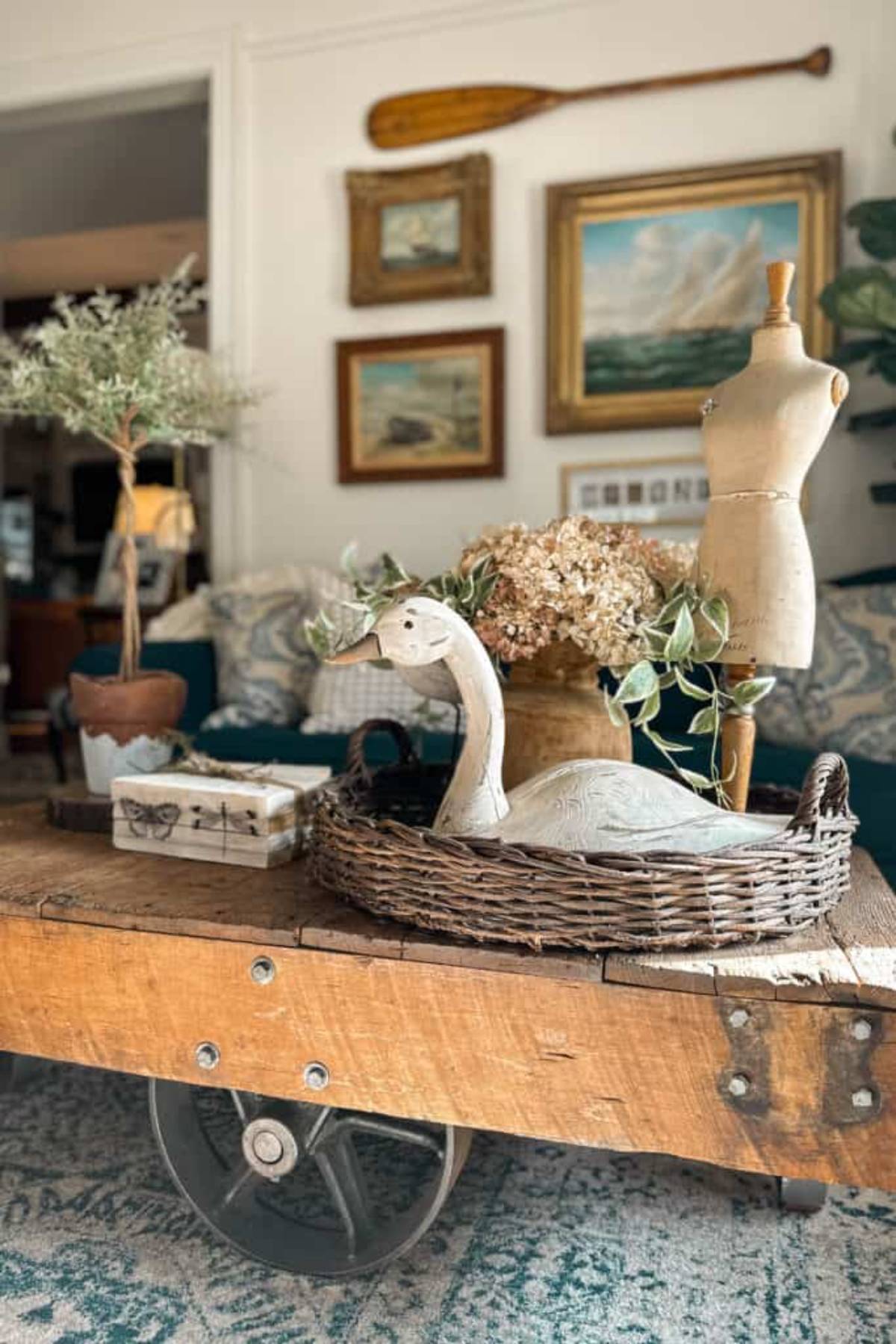 A cozy living room scene featuring a wooden cart coffee table with a wicker basket holding a ceramic duck, dried flowers, and greenery. In the background, a mannequin and framed paintings adorn the walls.