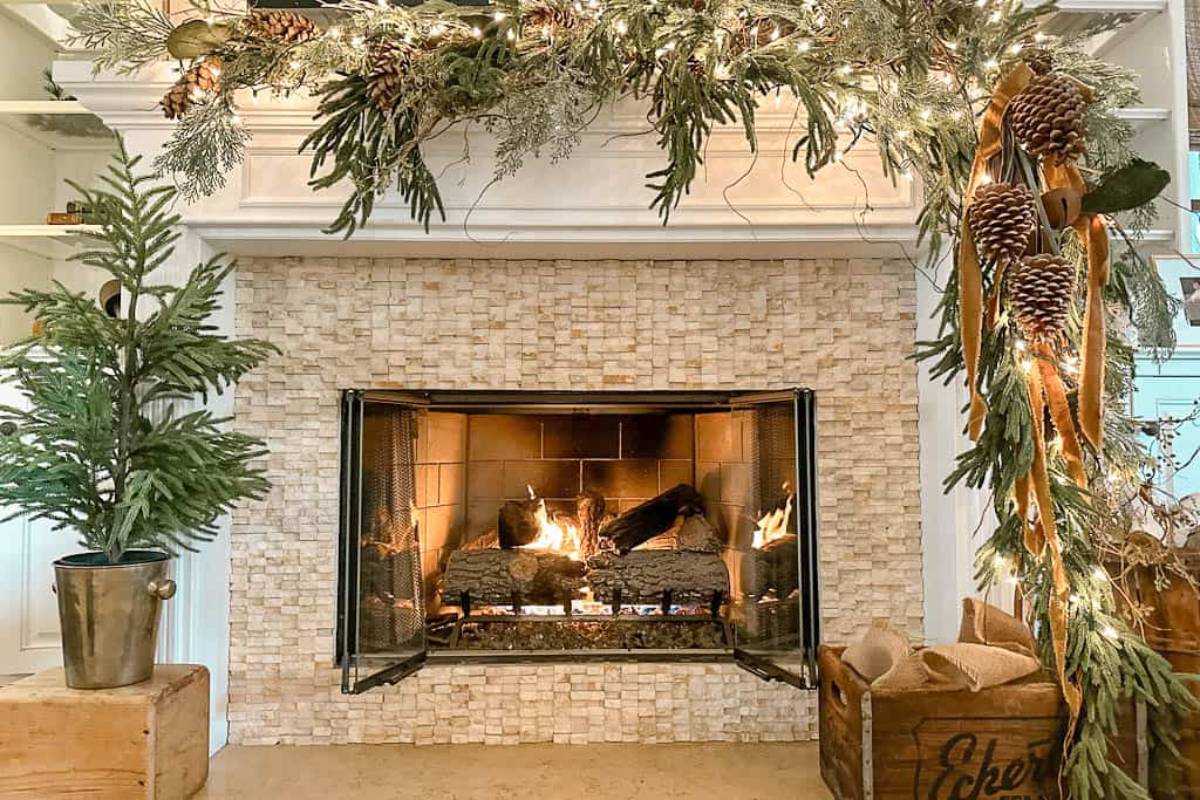 A cozy fireplace decorated with greenery, pine cones, and warm lights. Logs are burning, surrounded by a stone facade. A small spruce in a bucket and festive garland adorn the mantel. A burlap sack sits nearby.