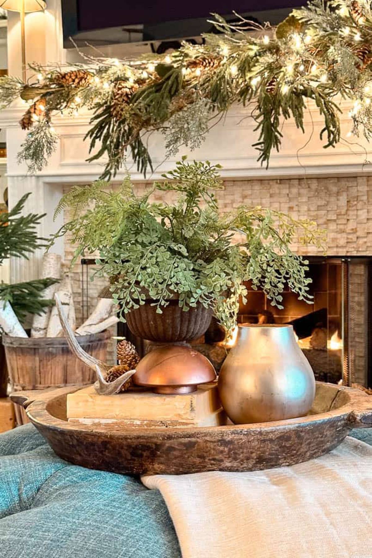 A cozy living room scene featuring a wooden tray with a green potted plant, a metallic teardrop-shaped vase, and decorative items. In the background, a fireplace is adorned with twinkling lights and greenery, creating a warm, inviting atmosphere.