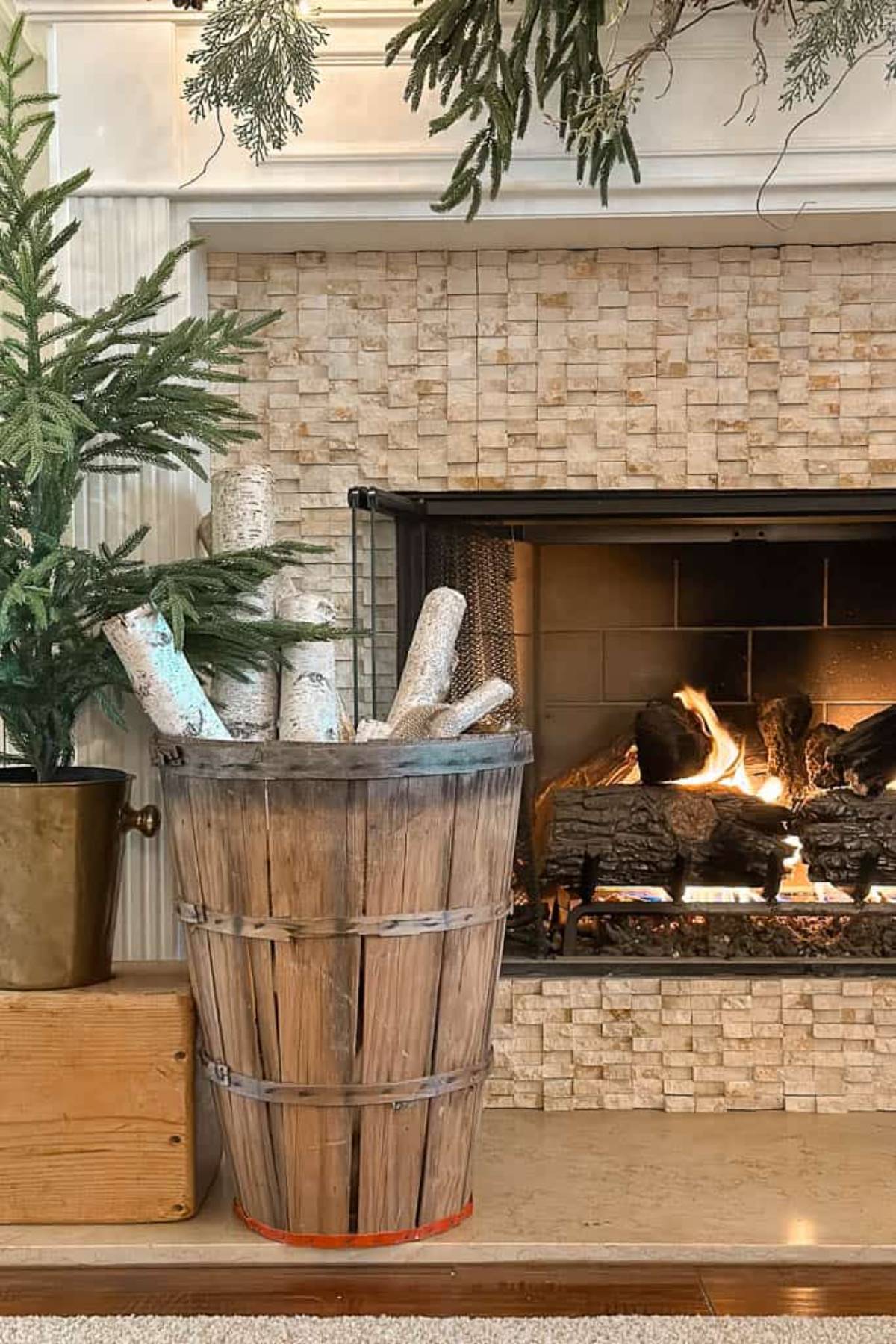 A cozy fireplace with logs burning, surrounded by a stone facade. A wooden basket filled with birch logs stands to the left, next to a potted evergreen plant. Greenery is draped above the fireplace, adding a festive touch.