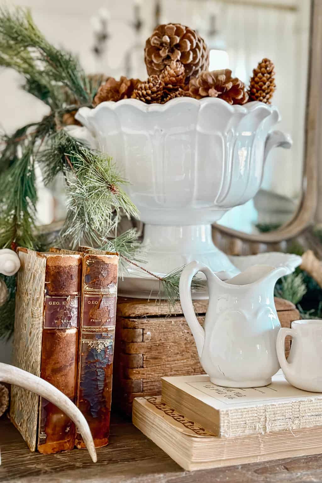 In a charming She Shed setting, a vintage arrangement features a white vase filled with pinecones, a white ceramic pitcher, and old books. Evergreen branches add to the rustic appeal, while the scene is beautifully reflected in a mirror—perfect for Sunday Sharing inspirations.