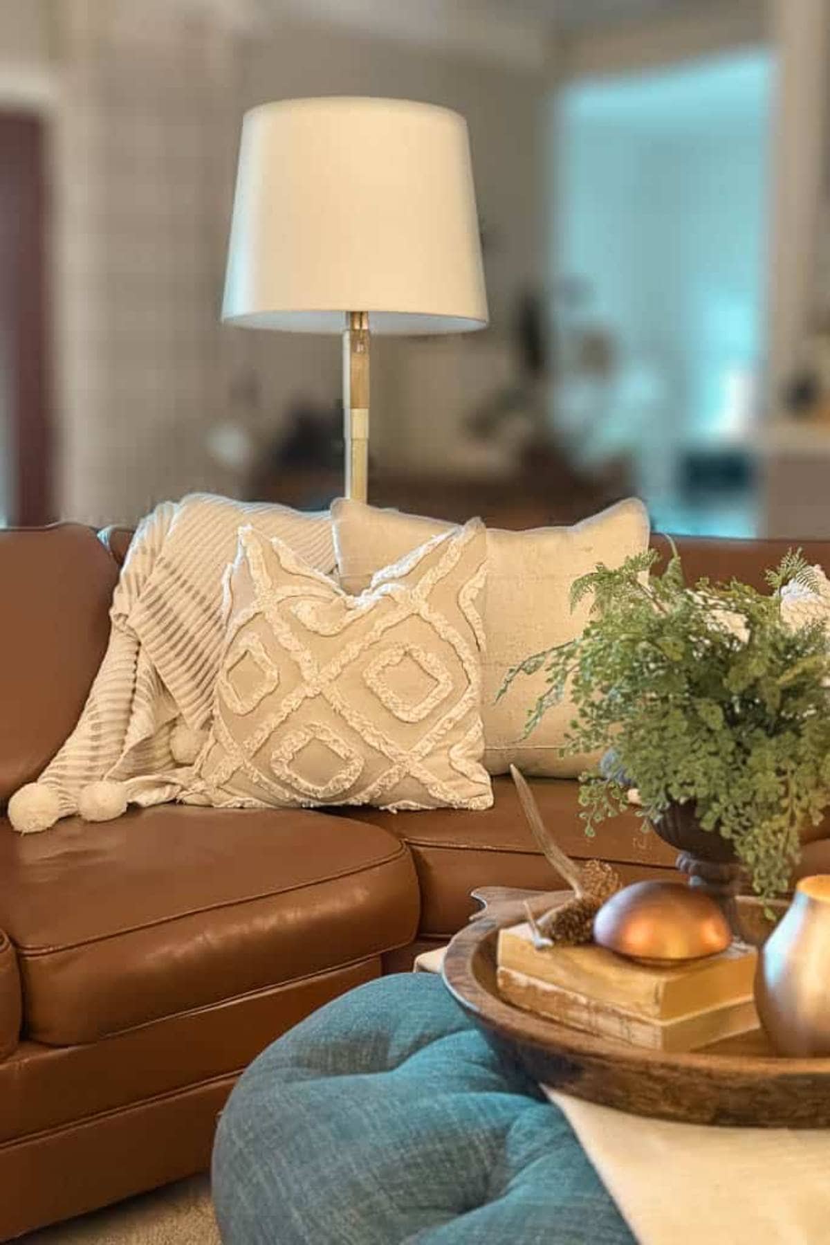 A cozy living room scene featuring a brown leather sofa with beige patterned cushions and a white throw. A floor lamp with a white shade stands behind the sofa. A wooden tray with decor items is on an upholstered blue ottoman.