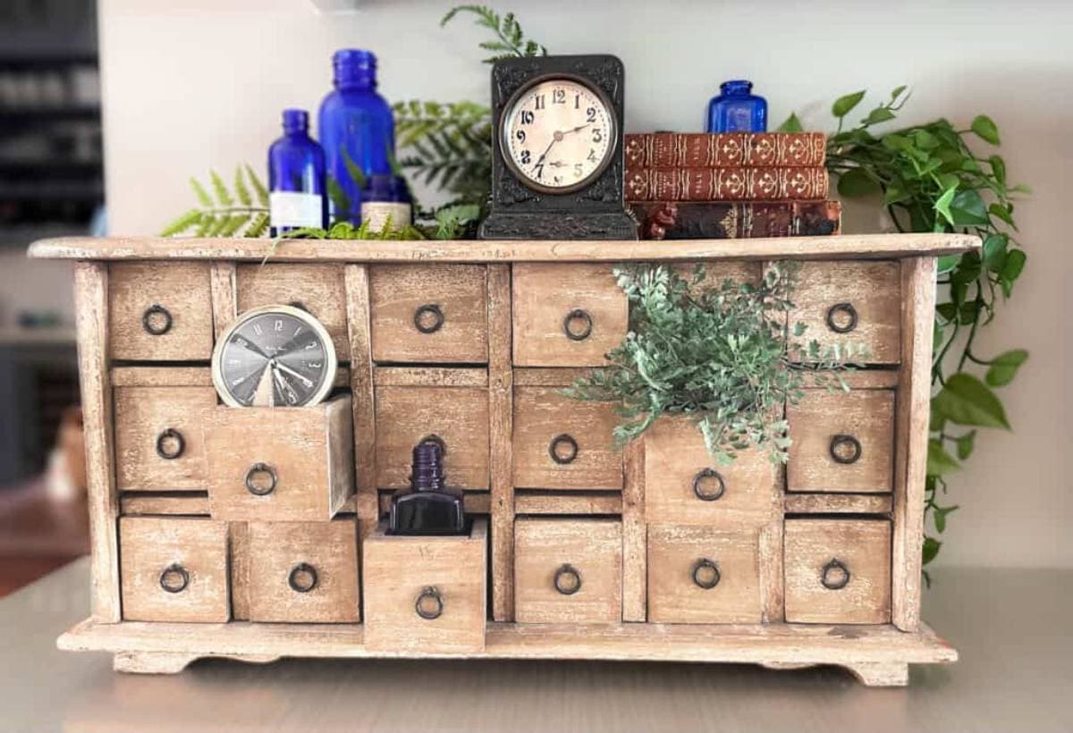 A wooden cabinet with multiple small drawers, some open, displaying items like a pocket watch, blue bottles, books, and greenery. A vintage clock and more plants sit on top, creating a rustic, antique look.