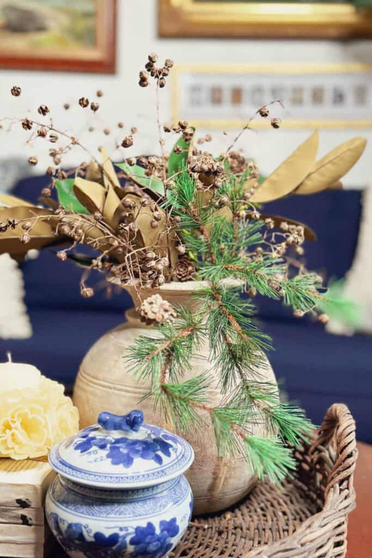 A ceramic vase with dried foliage and pine branches sits on a wicker tray alongside a blue and white porcelain jar with a lid, featuring floral patterns. A carved yellow candle shaped like a flower is next to them. A navy sofa is in the background.