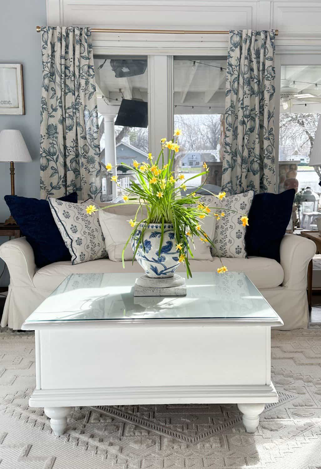 A cozy living room, perfect for Sunday Sharing, features a white sofa adorned with patterned blue and white cushions. A white and blue vase with yellow flowers graces the glass-topped coffee table. Blue floral curtains frame the large windows, allowing in natural light.