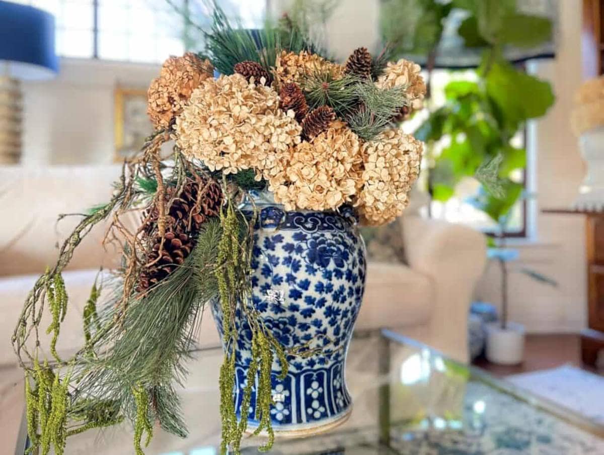 A blue and white ceramic vase holds a floral arrangement with dried hydrangeas, pinecones, and greenery. The vase sits on a glass coffee table in a bright living room with a beige couch and leafy plants in the background.