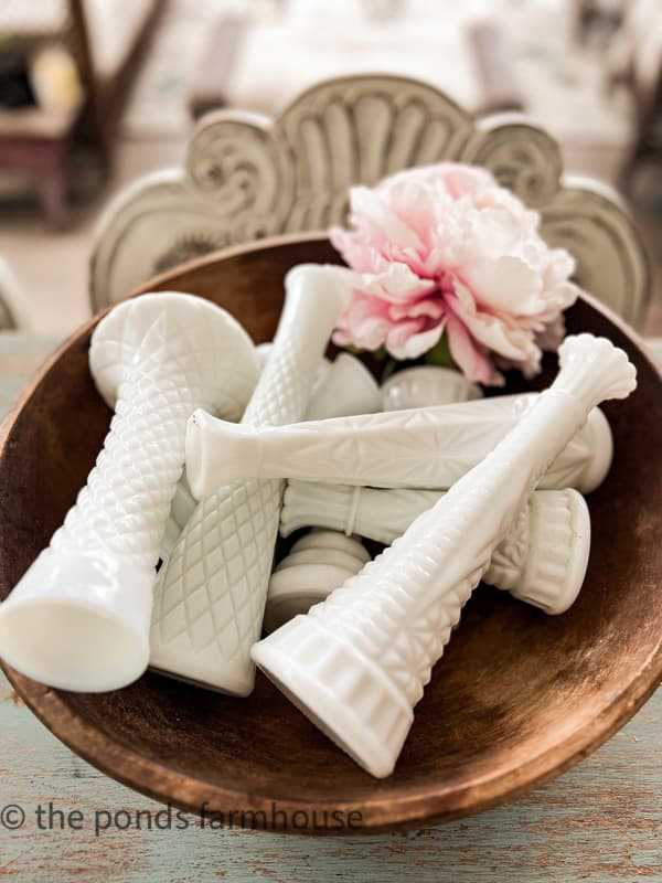 In a cozy She Shed setting, a wooden bowl cradles several white, intricately patterned vase bases with a pink peony softly resting on top. The backdrop hints at nostalgia with a blurred, vintage-style chair. Perfect for Sunday Sharing moments.