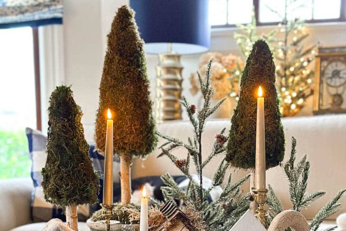 A cozy living room decorated for the holidays, featuring small evergreen tree sculptures and lit candles on a table. In the background, a lamp, a window, and a Christmas tree with lights are visible.