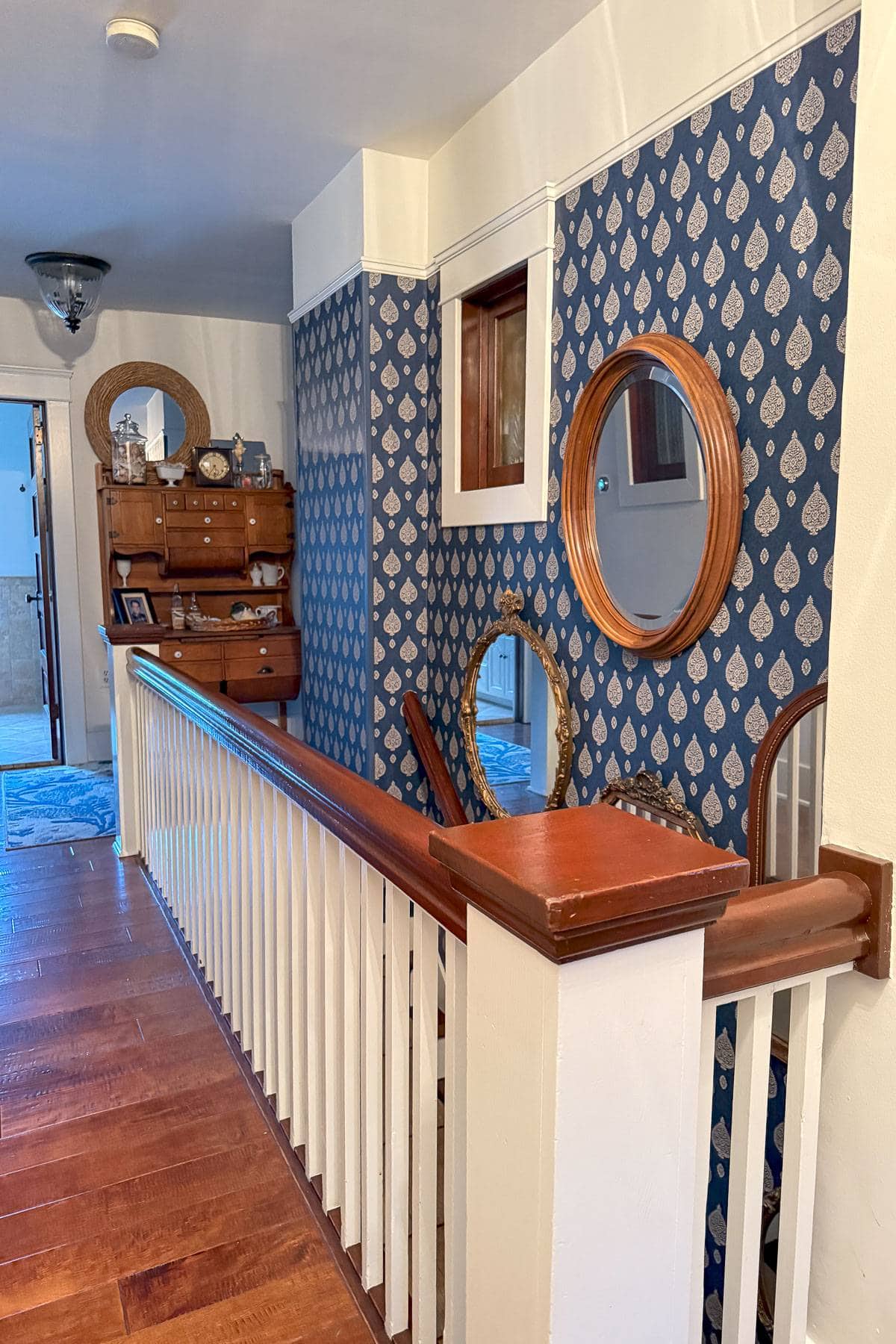 Hallway with wooden flooring and white railing, featuring a blue patterned accent wall. Three round mirrors and decorative plates adorn the wall. A wooden cabinet with shelves and various items is visible in the corner.