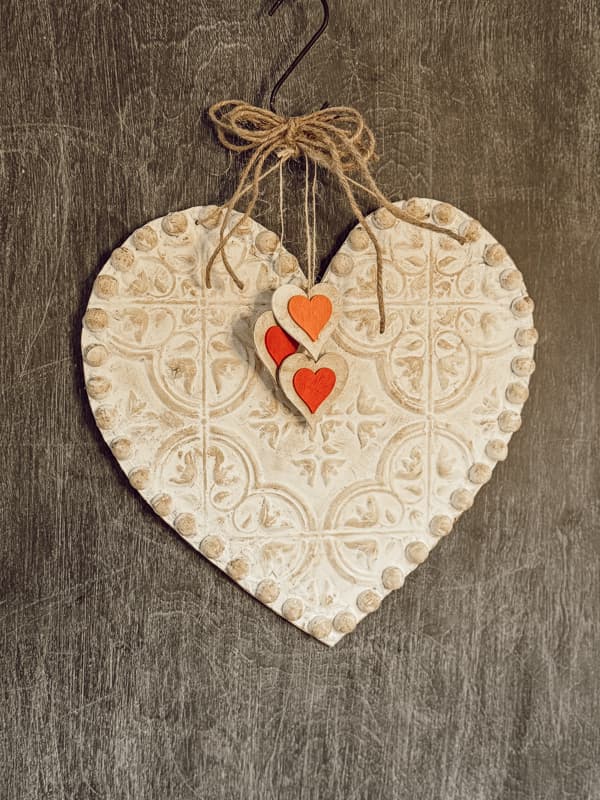 Decorative heart-shaped ornament with an intricate pattern, adorned with two smaller red hearts, hangs by a twine bow in a cozy She Shed against a textured gray background.