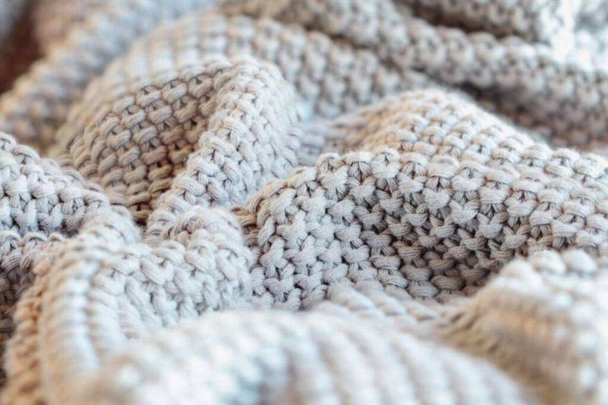 Close-up of a textured, woven gray blanket with a chunky knit pattern. The soft, undulating surface creates a cozy and inviting appearance, with subtle shadows enhancing the depth and detail of the fabric.