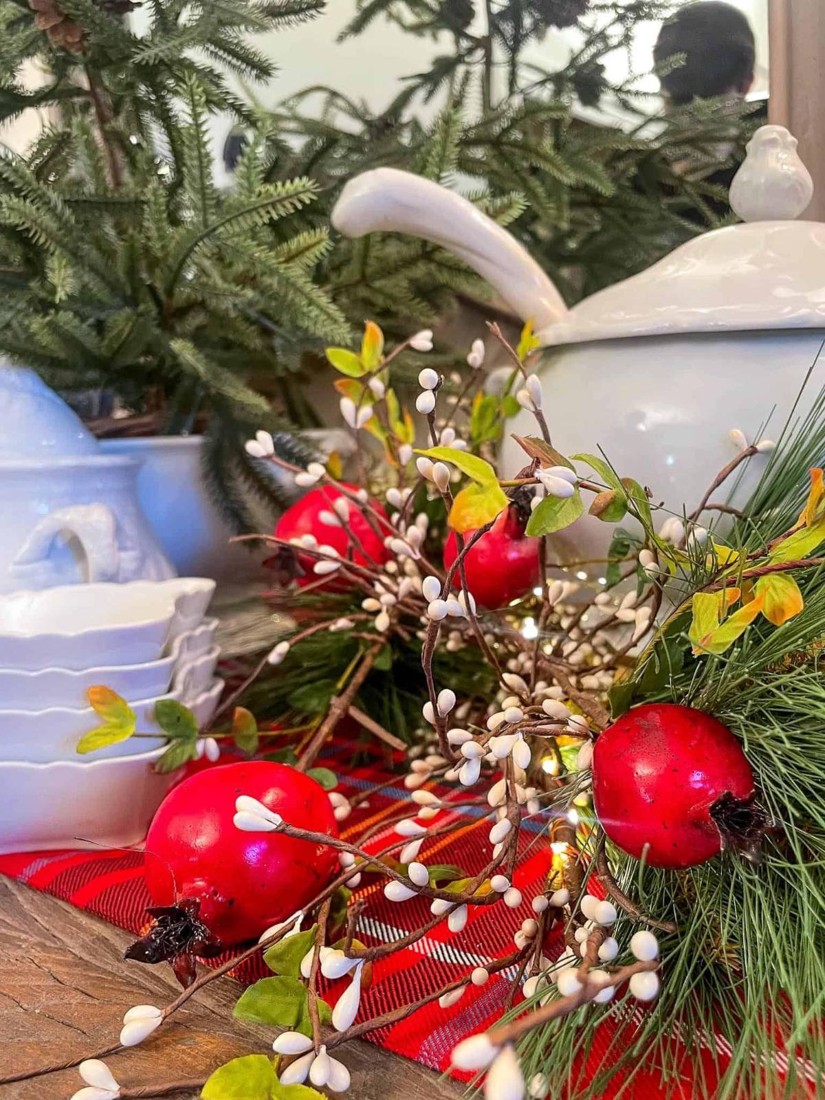 A festive tabletop display featuring white ceramic dishes and a lidded pot surrounded by green pine branches, red apples, and white berry sprigs on a red checkered cloth.