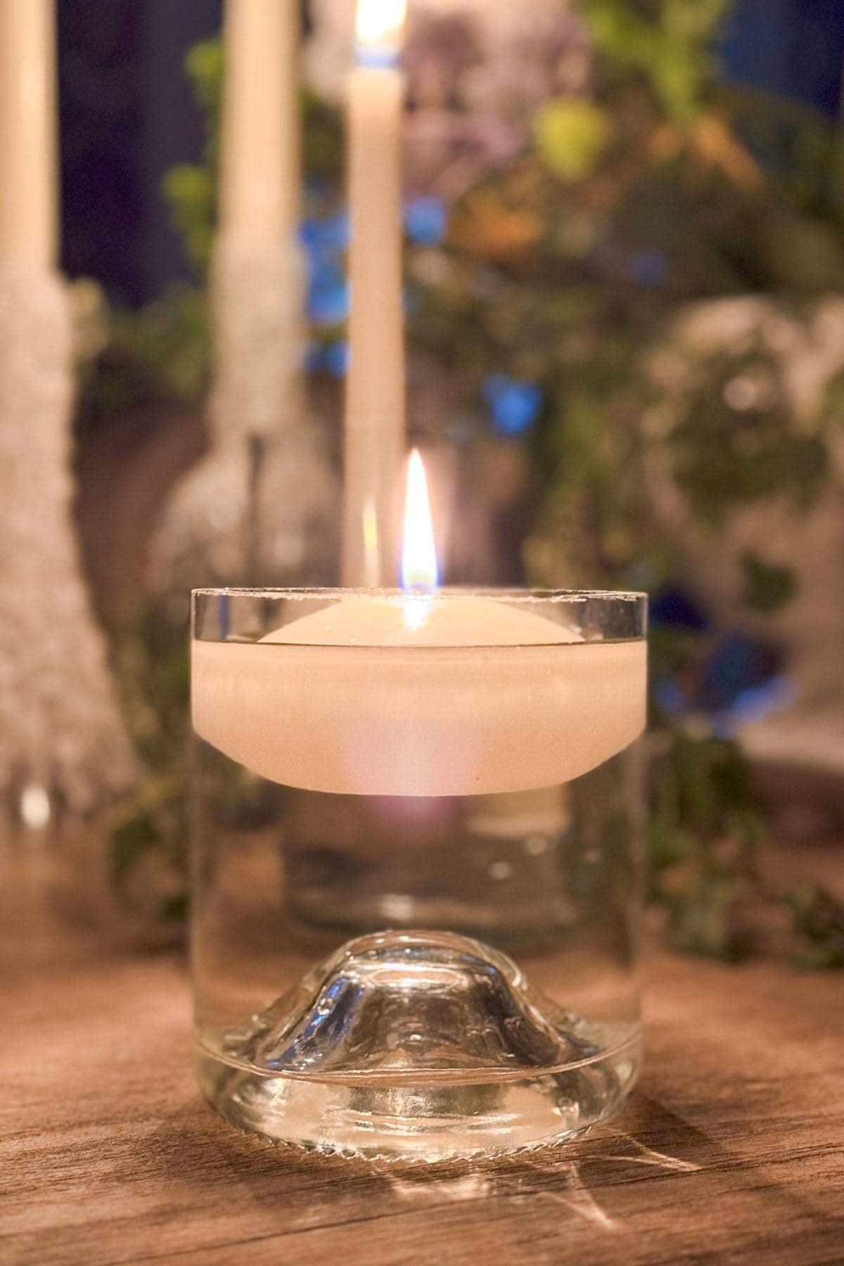 A white floating candle is lit in a clear glass filled with water. The scene is warm and inviting with blurred greenery and additional candles in the background on a wooden surface.