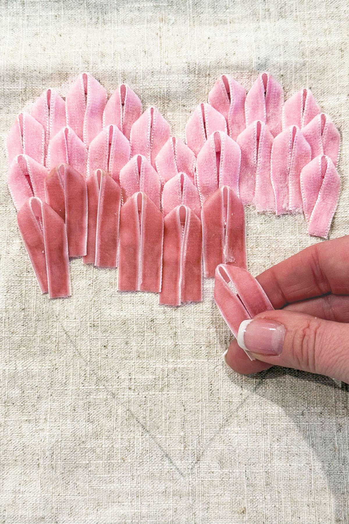 A hand placing a pastel pink fabric petal onto a heart shape made of similar petals on a beige fabric surface. The petals are neatly arranged to form the shape of a heart.