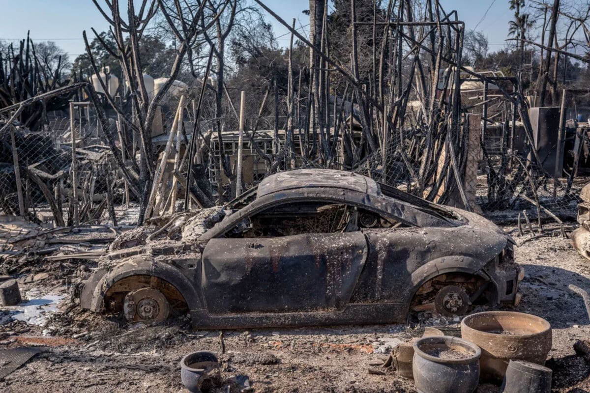 A burned-out car sits amid the charred remains of a destroyed landscape, surrounded by twisted metal and debris. The scene, reminiscent of a chaotic Volume 33 out of an intense series, shows devastation likely caused by a fire, with trees and structures heavily damaged in the background.