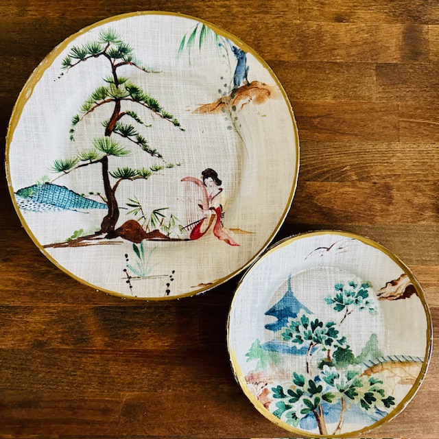 Two decorative plates with intricate designs. The larger plate features a figure near a tree and rocks, while the smaller plate depicts a landscape with trees and a mountain. Both plates have an earthy color palette and are set on a wooden surface.